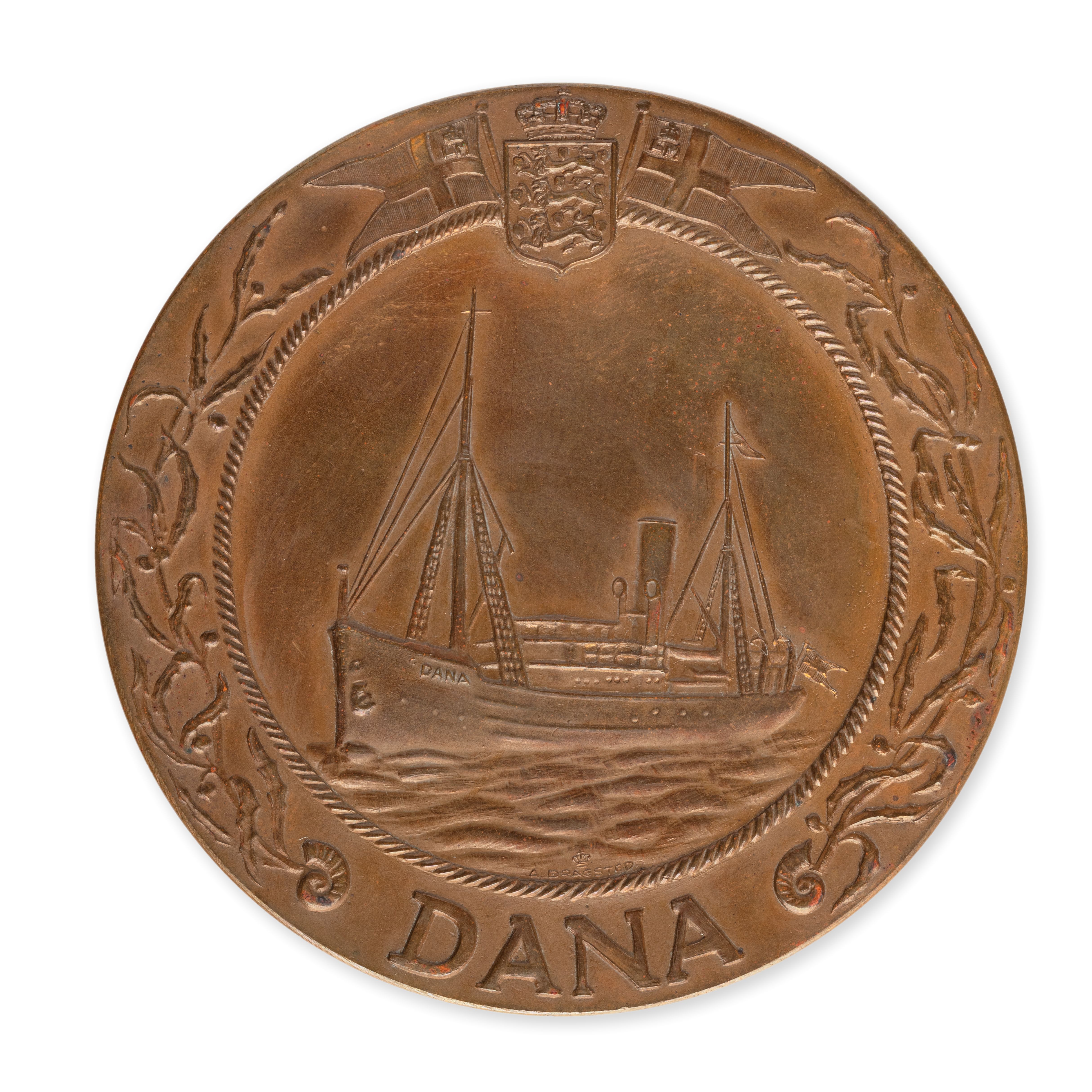 Bronze-like medal engraved with the picture of an ocean vessel with two masts, a funnel and with the name ‘Dana’ on its bow and at the bottom of the medal.