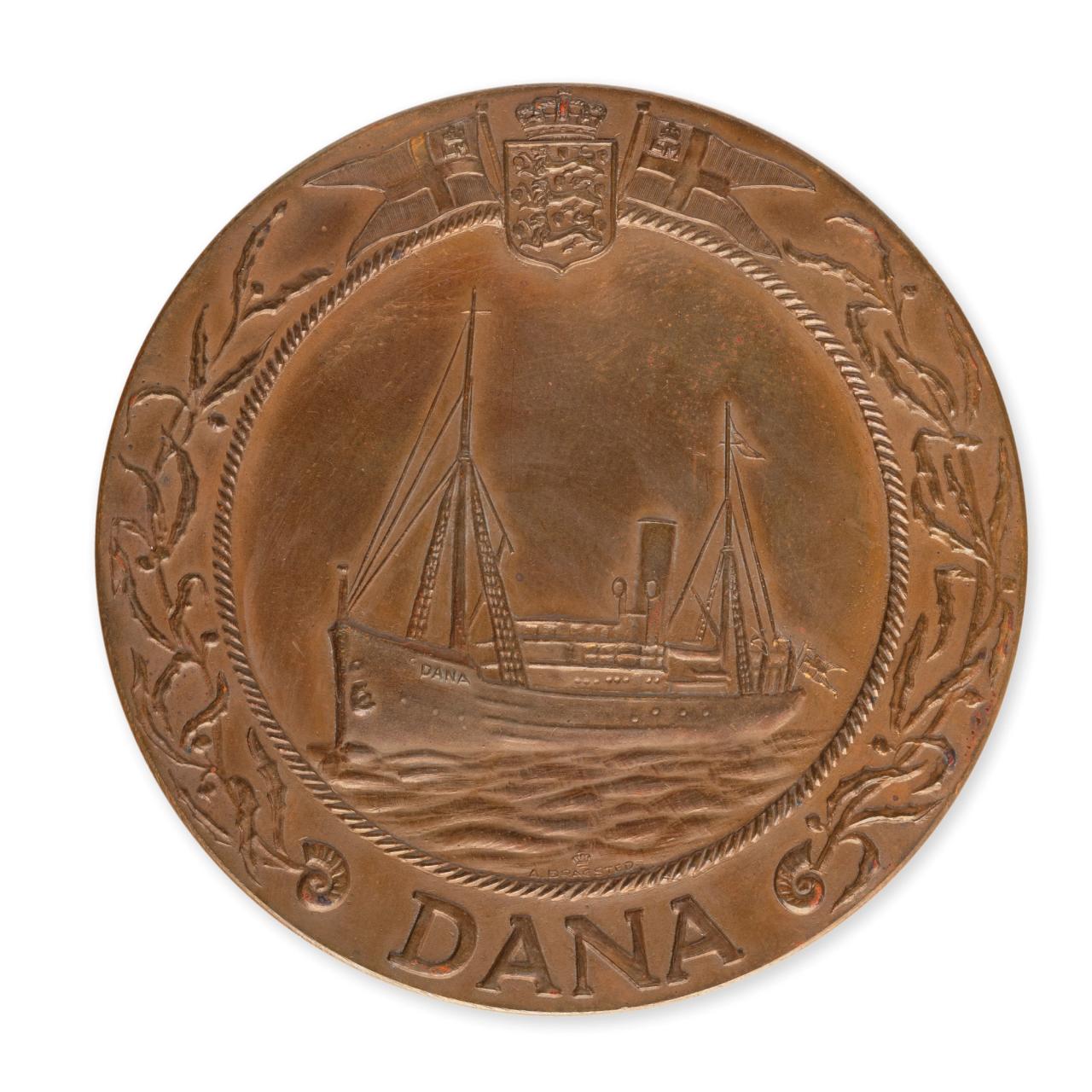 Bronze-like medal engraved with the picture of an ocean vessel with two masts, a funnel and with the name ‘Dana’ on its bow and at the bottom of the medal.
