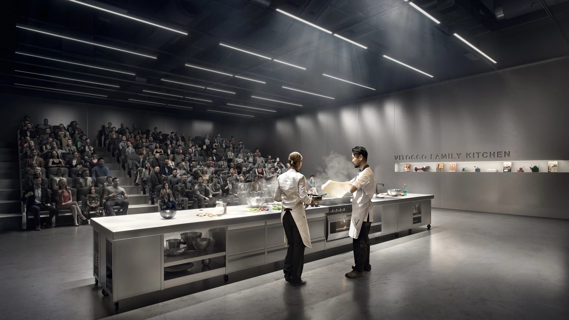 Two figures are cooking at a long silver kitchen island. An audience are watching, sitting in theatre seating.