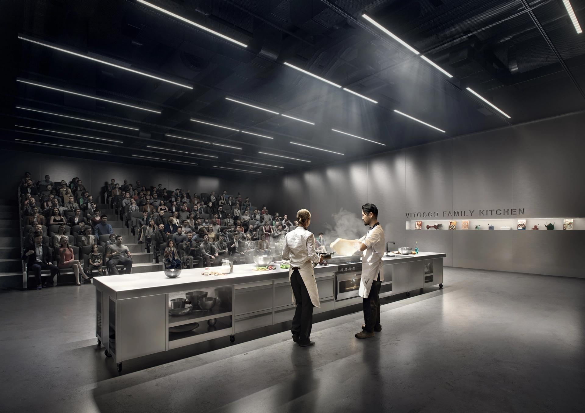 Two figures are cooking at a long silver kitchen island. An audience are watching, sitting in theatre seating.