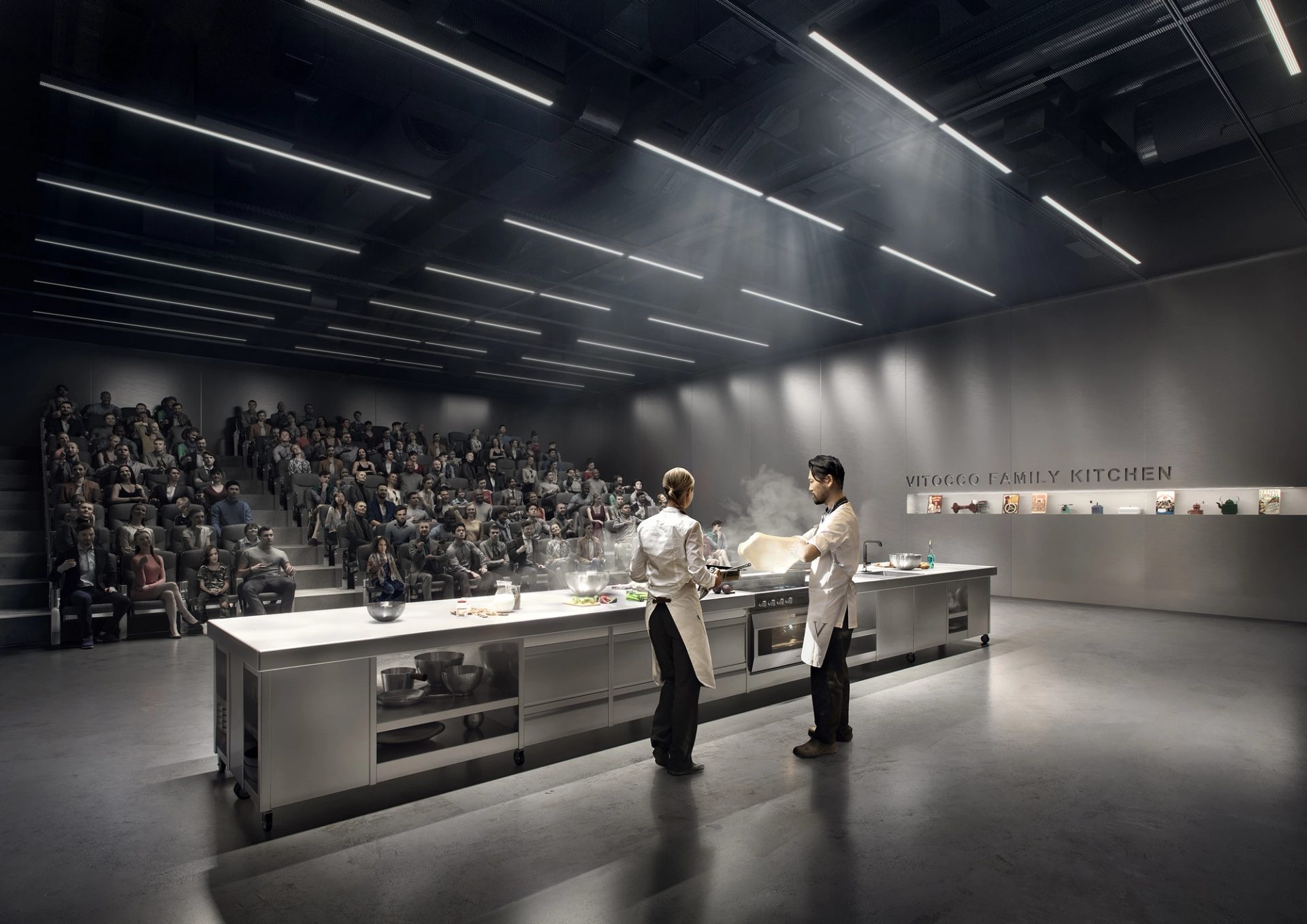 Two figures are cooking at a long silver kitchen island. An audience are watching, sitting in theatre seating.