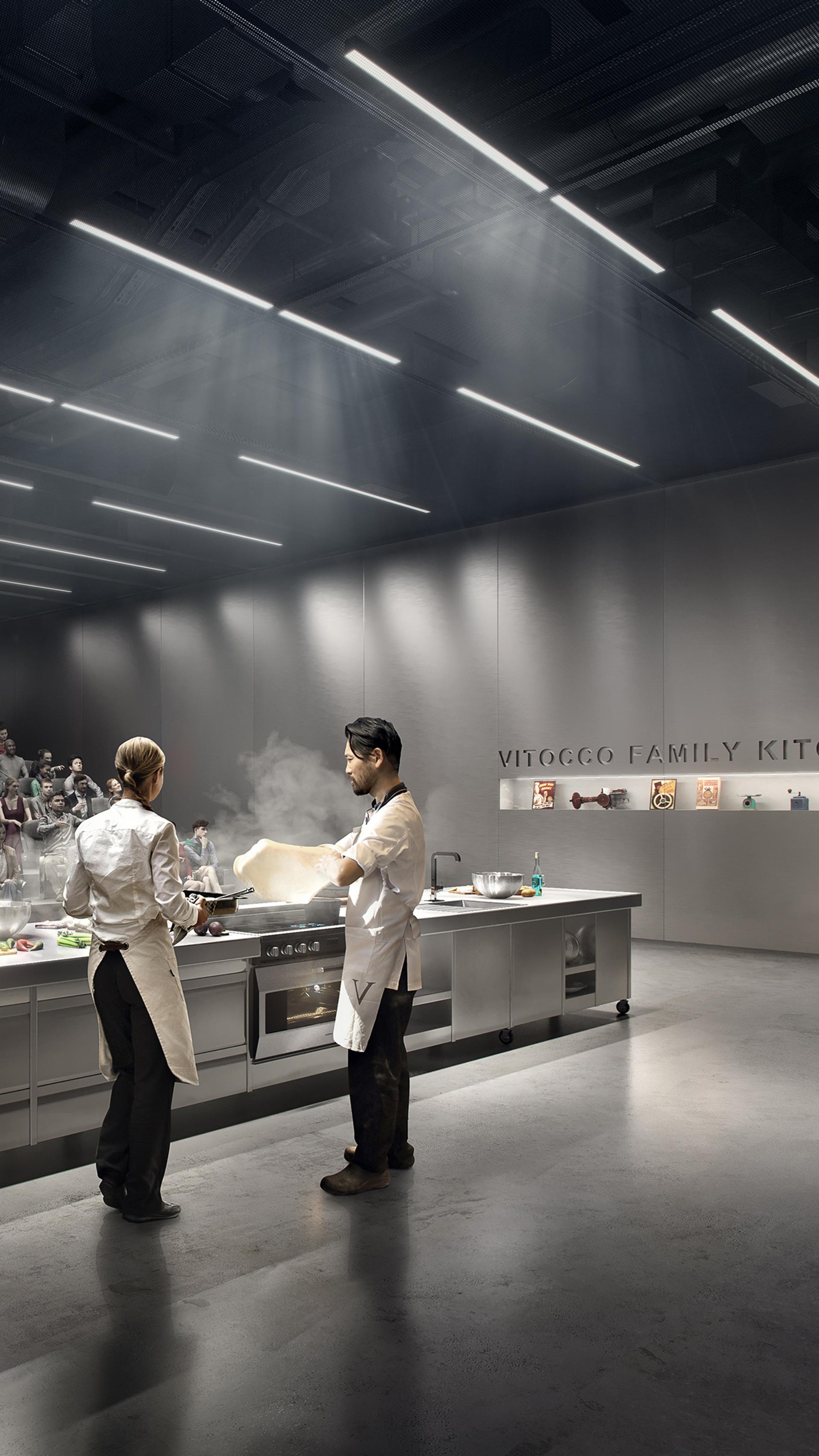 Two figures are cooking at a long silver kitchen island. An audience are watching, sitting in theatre seating.