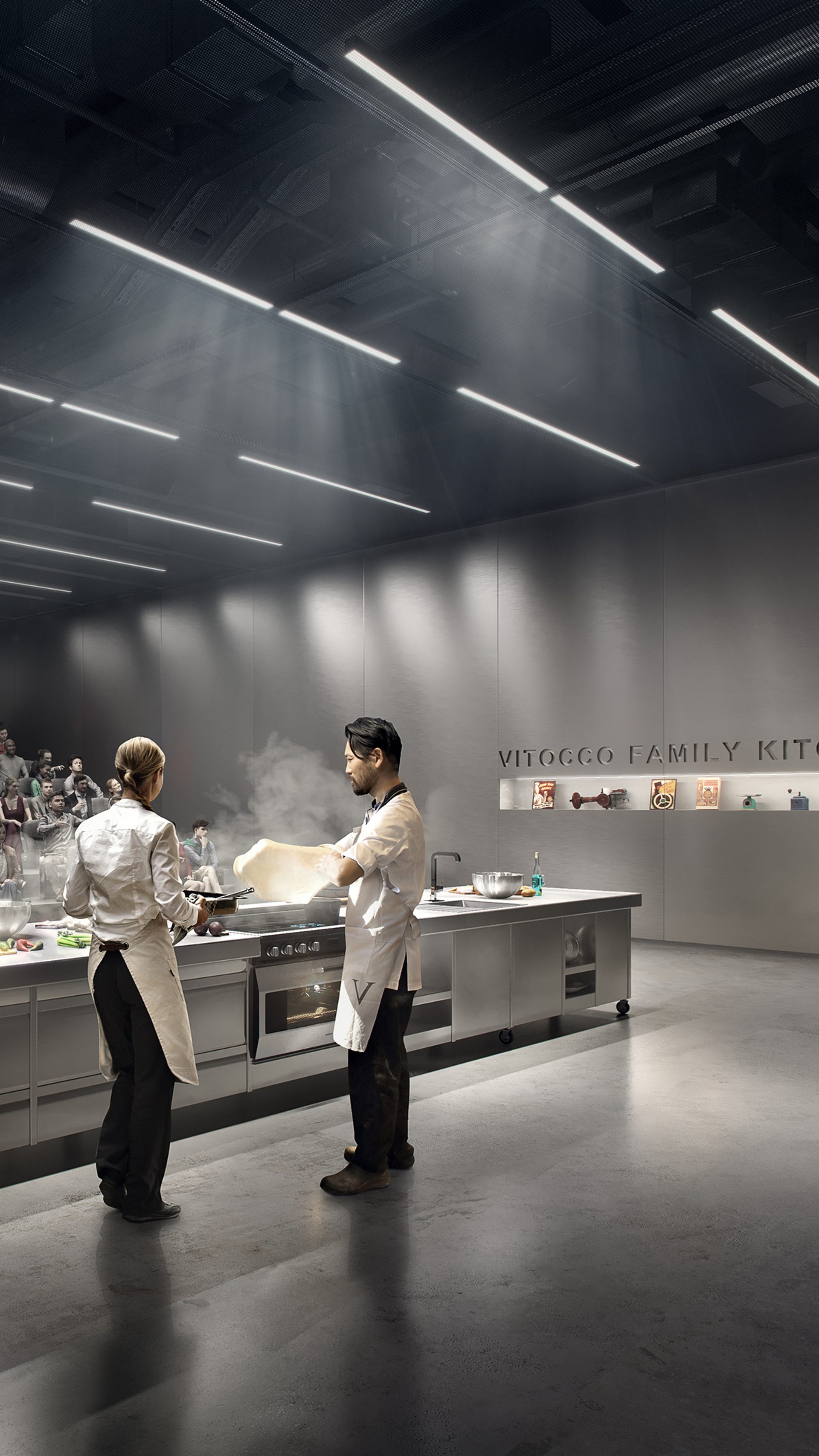 Two figures are cooking at a long silver kitchen island. An audience are watching, sitting in theatre seating.