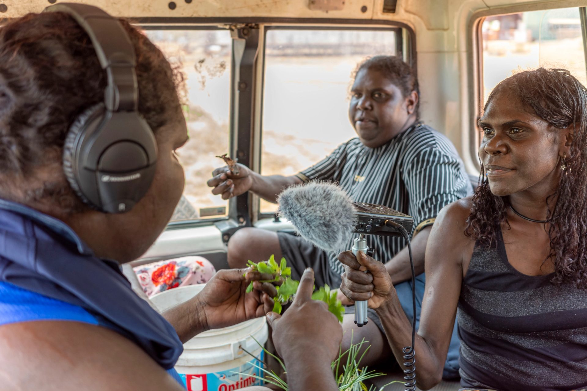 Three women sitting together in the back of a troopie. The woman at the top right of the frame is holding a microphone and the woman at the top left of the frame is speaking into it. She is also holding green leaves in her right hand and wearing black over ear headphones. The woman at the back is holding a brown piece of plant in her right hand.