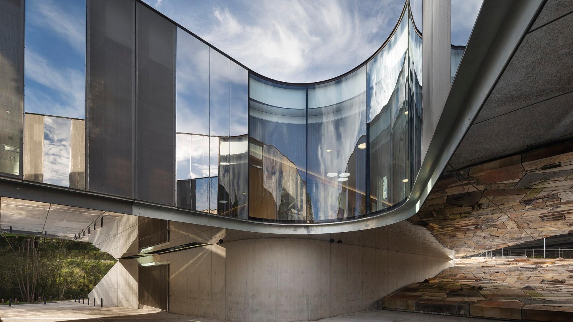 The curving cement, steel and glass exterior of the Australian PlantBank under a bright blue sky. The sky reflects on the building’s partially mirrored surfaces.