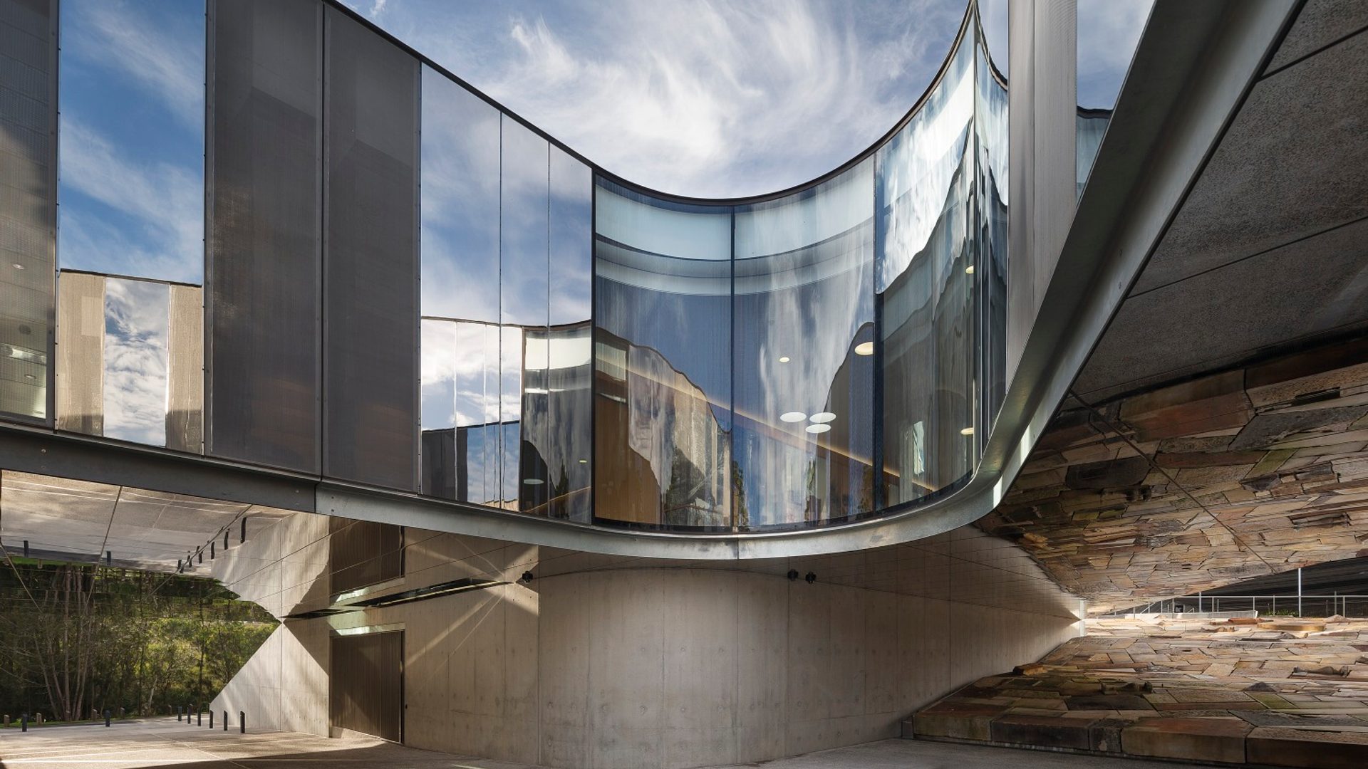 The curving cement, steel and glass exterior of the Australian PlantBank under a bright blue sky. The sky reflects on the building’s partially mirrored surfaces.