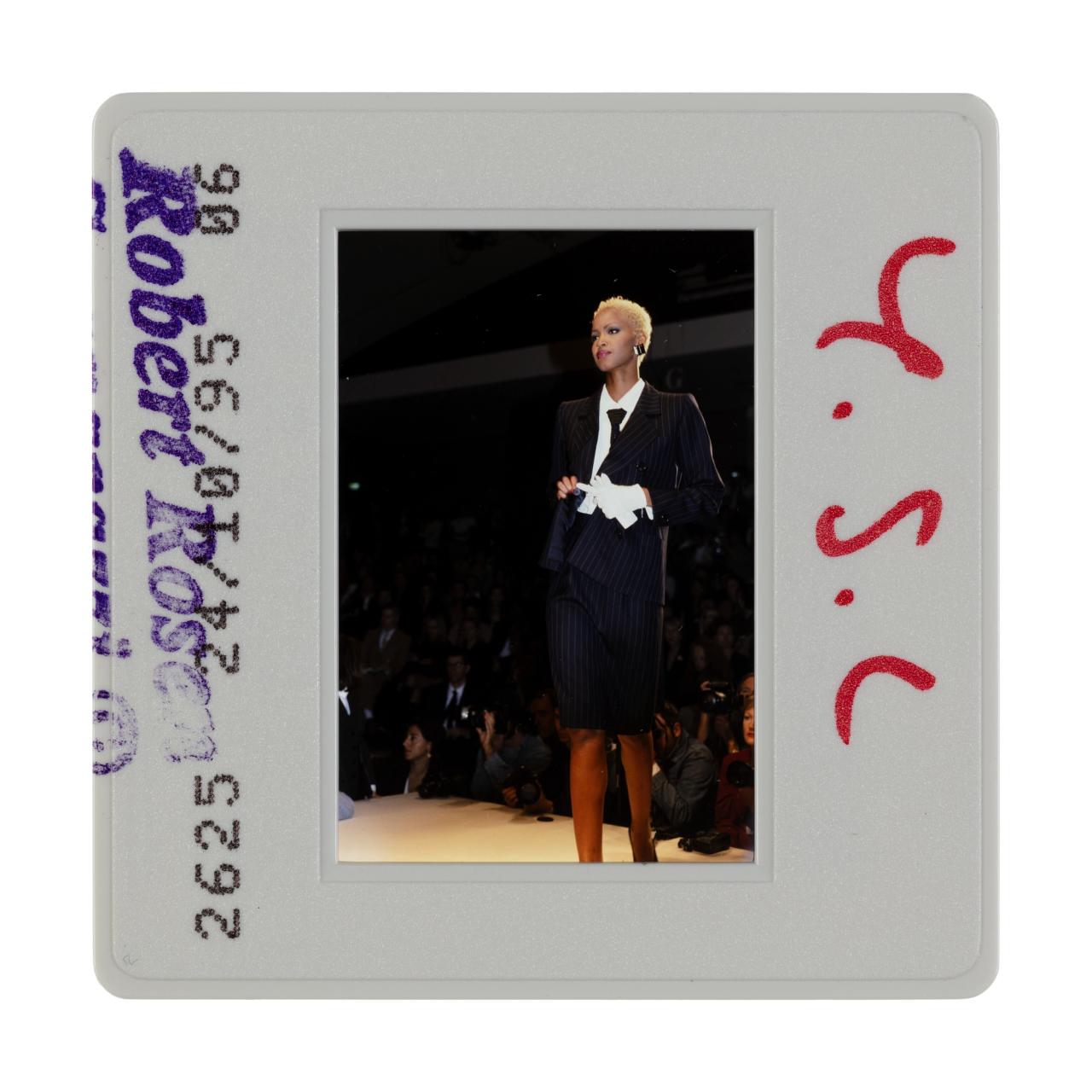 A photo positive 35mm slide, depicting a fashion show and runway model wearing a black pinstripe two piece suit jacket and pencil skirt, the model is removing their white gloves.