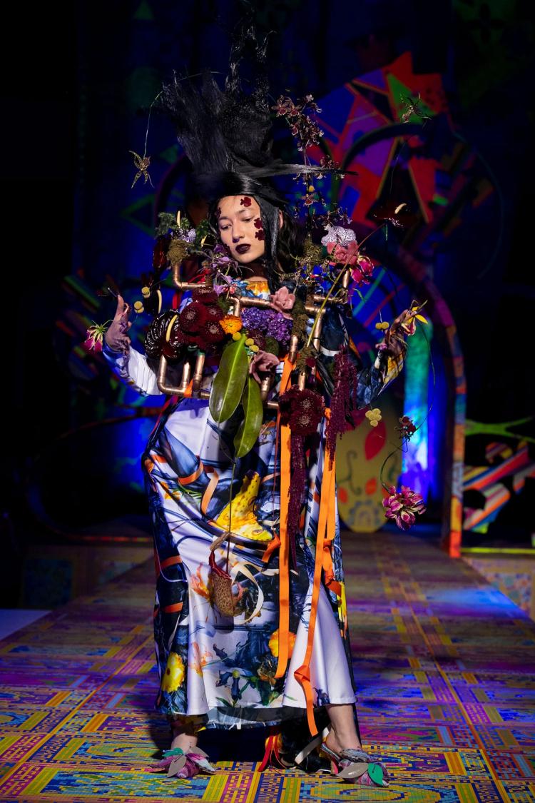 Runway model wearing a silk dress and copper pipes interlaced with flowers around their chest. On their head they are wearing a tall black pointed headpiece.