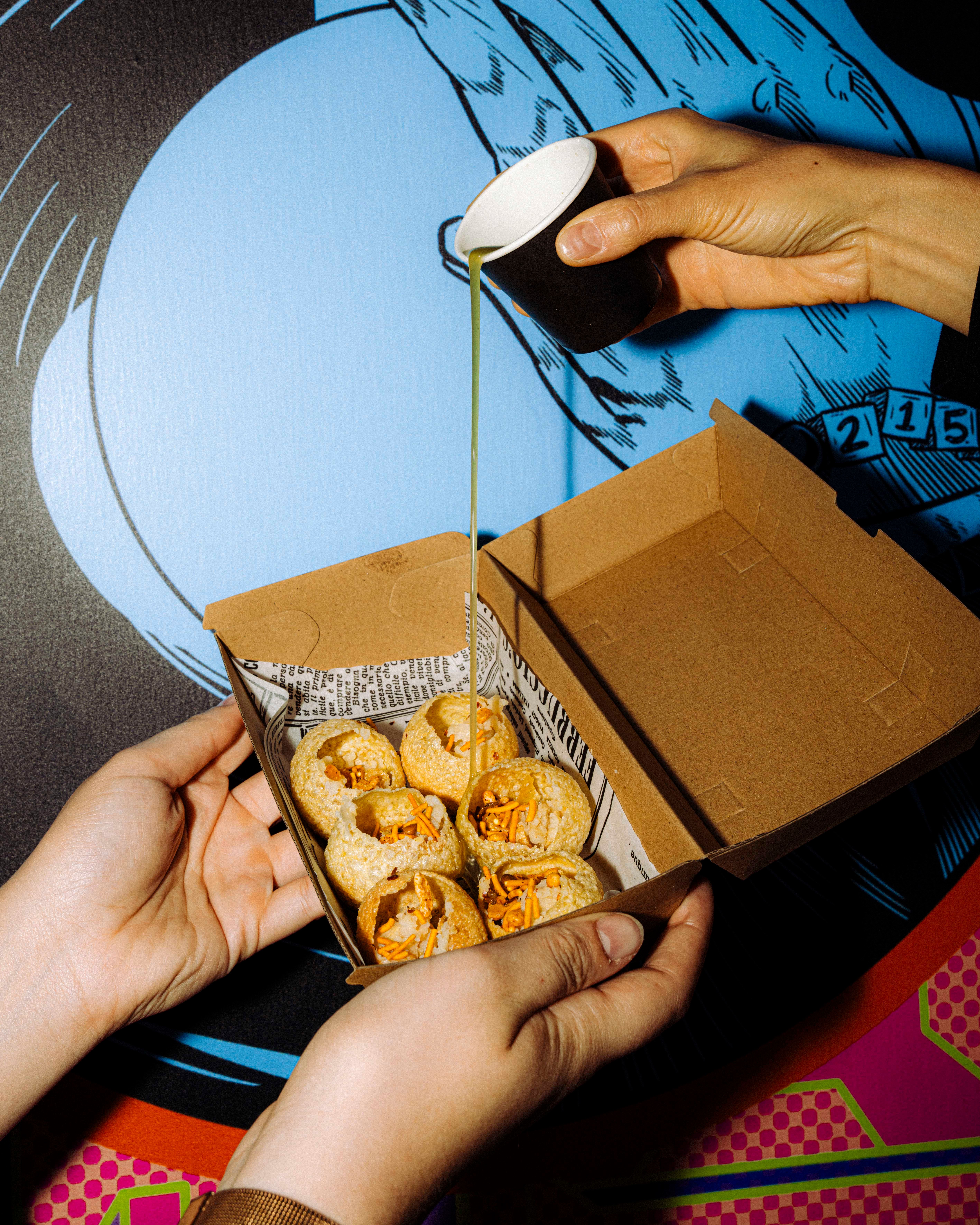 Two hands hold a cardboard box of round snacks filled with ingredients, while another hand pours green liquid from a small black cup onto the snacks.
