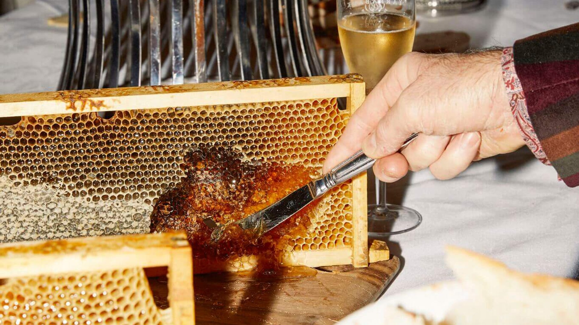 A table of various honeycombs and delicacies. A person uses a butter knife to scrape a small amount of dark brown honey from one section of honeycomb.