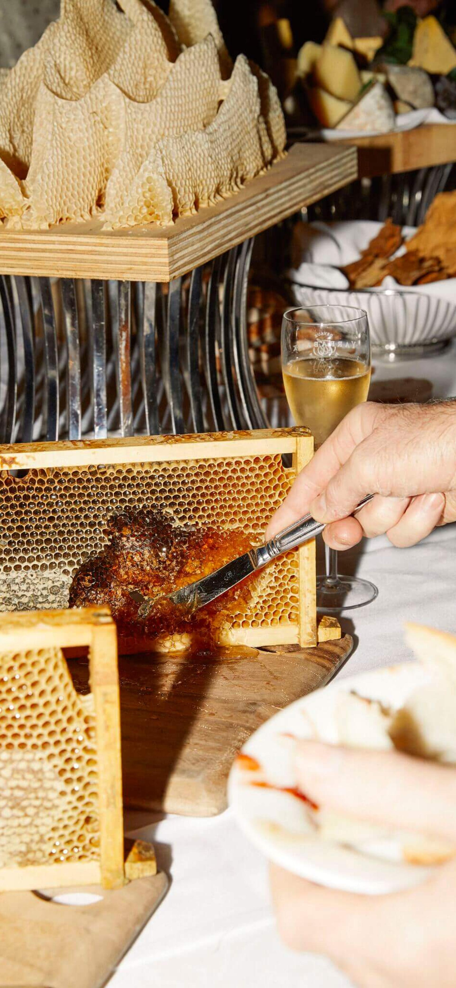 A table of various honeycombs and delicacies. A person uses a butter knife to scrape a small amount of dark brown honey from one section of honeycomb.