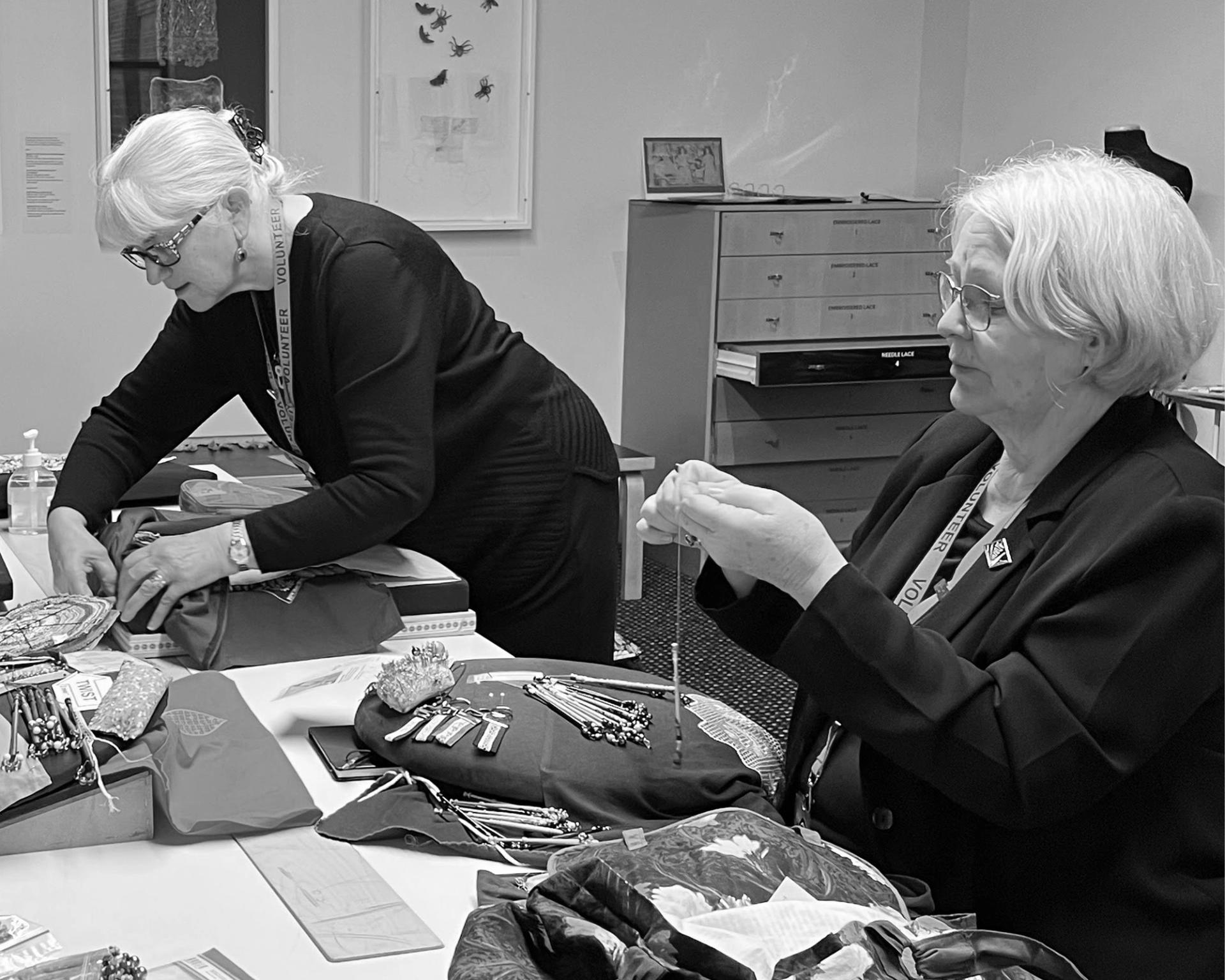 Black and white image of two women (left: unknown, right: Barbara Stokes) dressed in black and practicing lacemaking. They're using pillows and bobbin machines on a table in front of them to create lace.