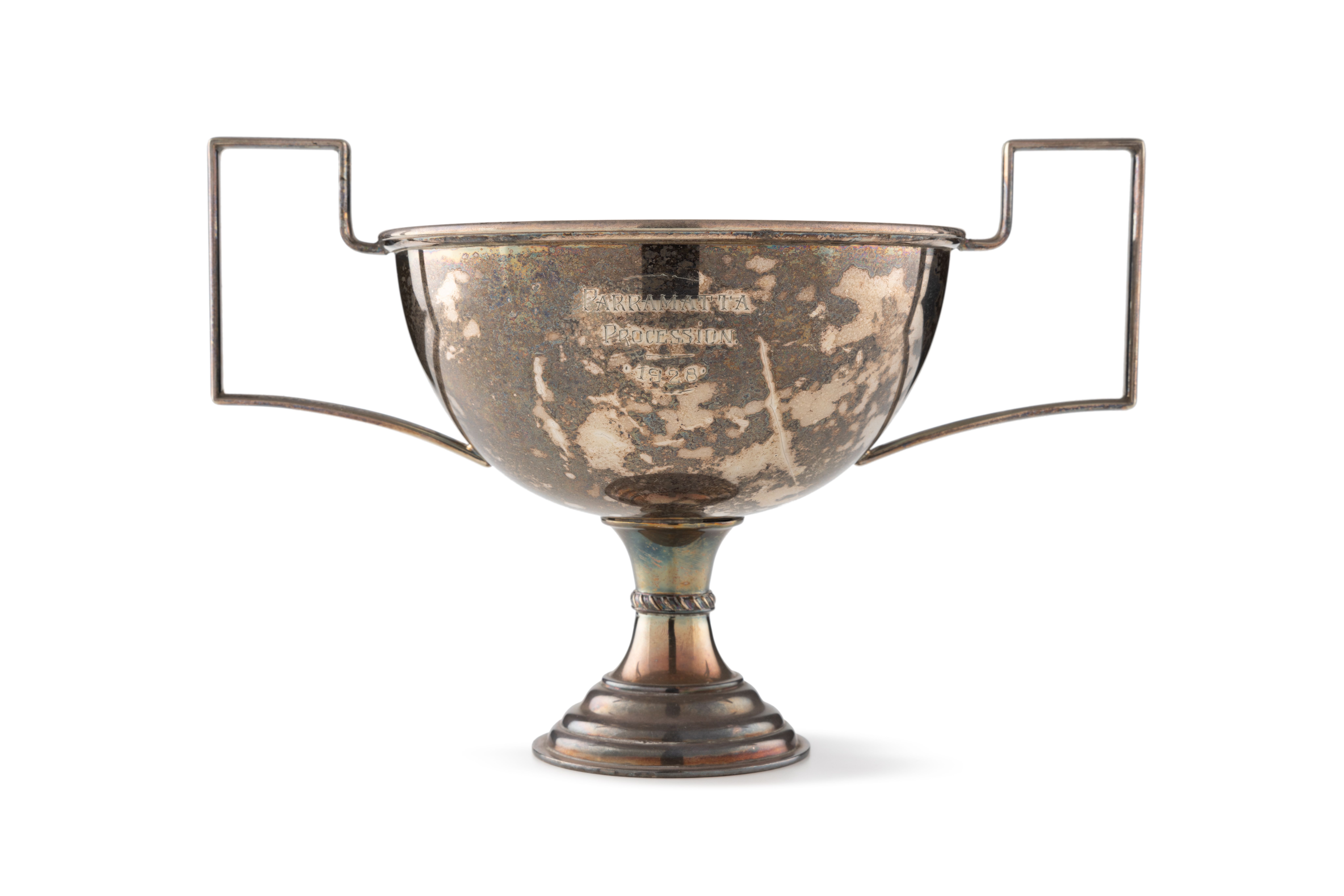 A metal trophy with a short, round bowl and intricate, angled handles on either side. Engraved on the front is 'Parramatta Procession, 1928'. The metal shows brown and light brown spots all over, with hints on blue shines along the curved base.