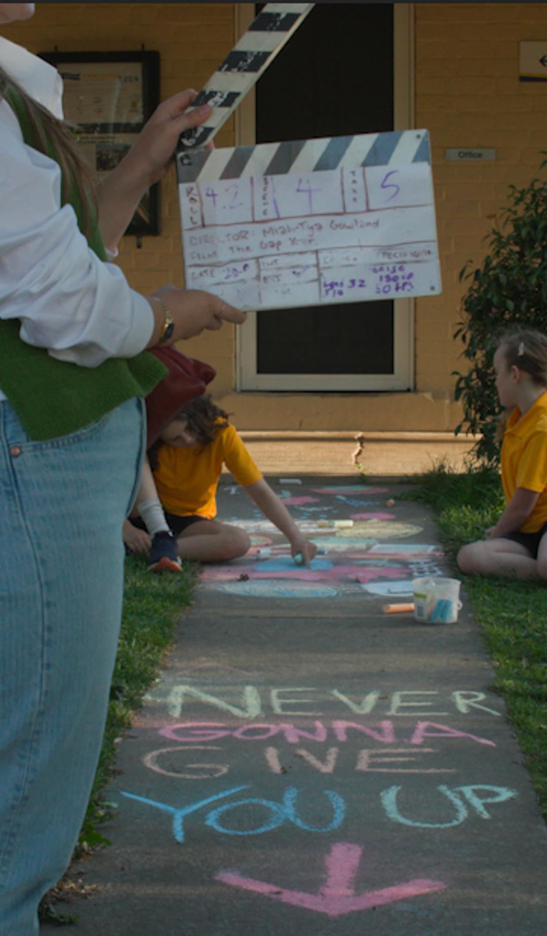 Crew member holding a clapper board with two young children in the background. Text written in chalk on the ground says "NEVER GONNA GIVE YOU UP"