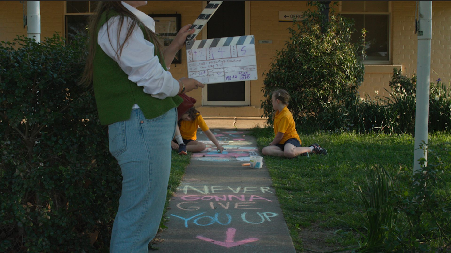 Crew member holding a clapper board with two young children in the background. Text written in chalk on the ground says "NEVER GONNA GIVE YOU UP"