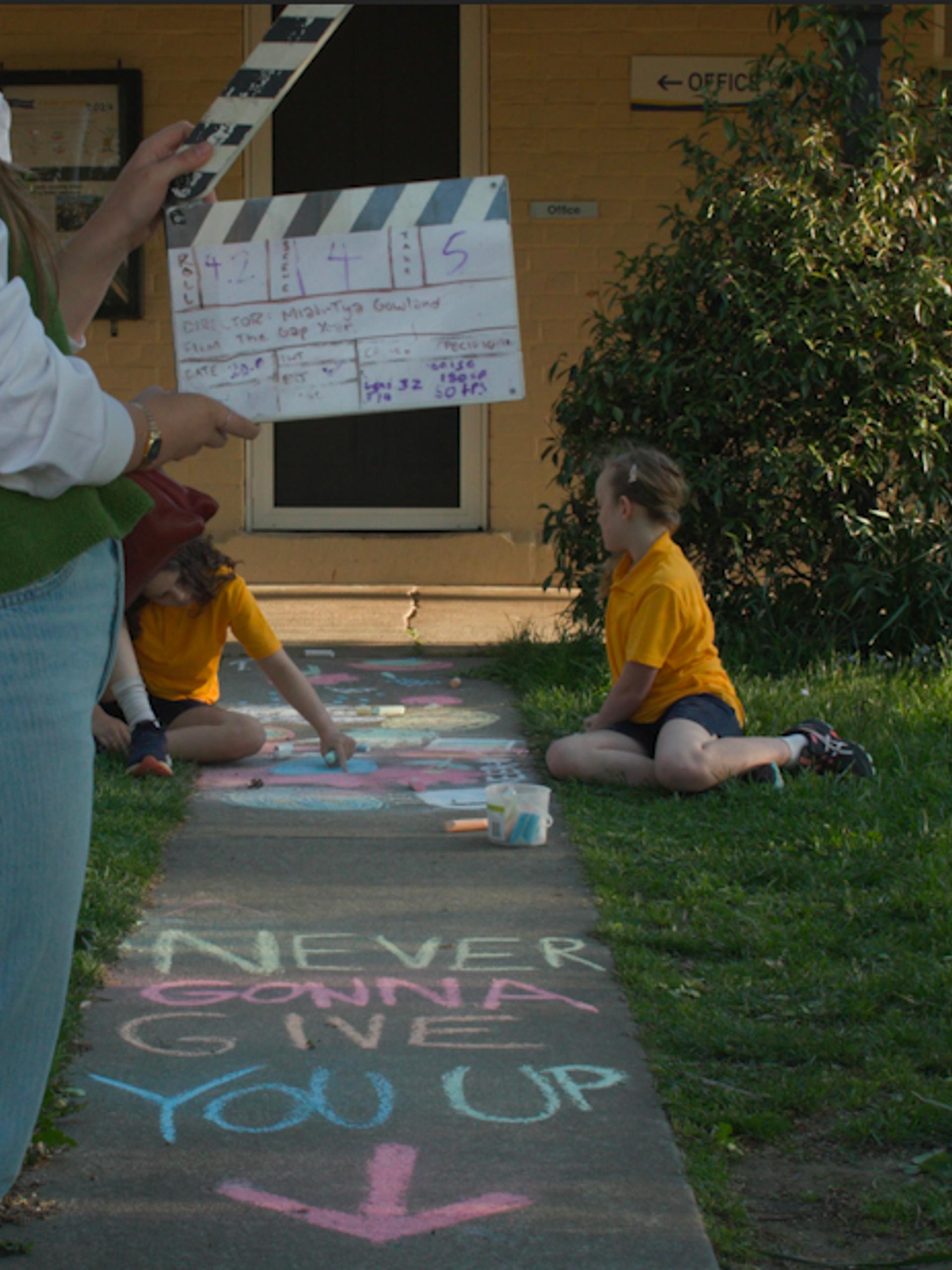 Crew member holding a clapper board with two young children in the background. Text written in chalk on the ground says "NEVER GONNA GIVE YOU UP"