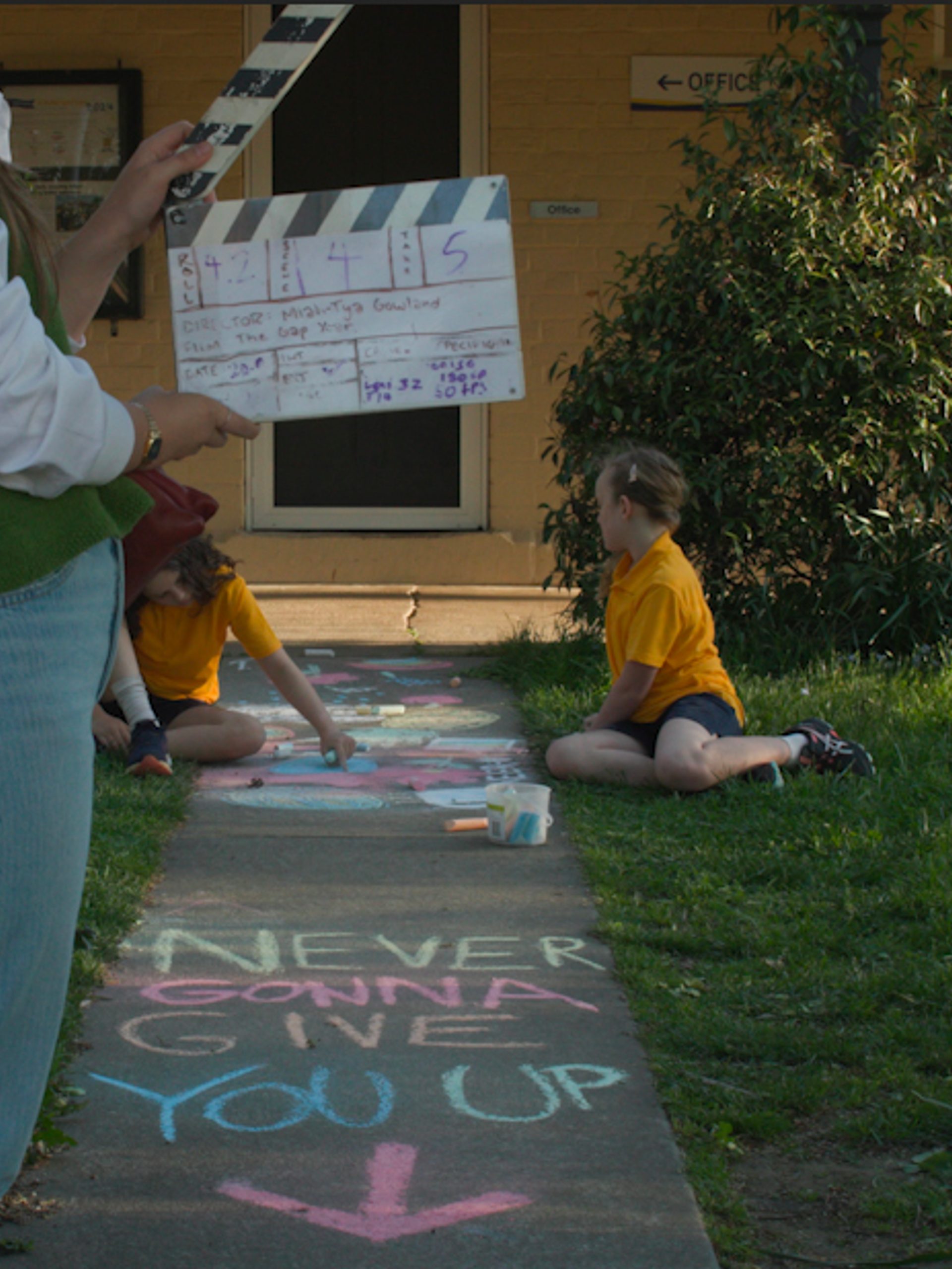 Crew member holding a clapper board with two young children in the background. Text written in chalk on the ground says "NEVER GONNA GIVE YOU UP"