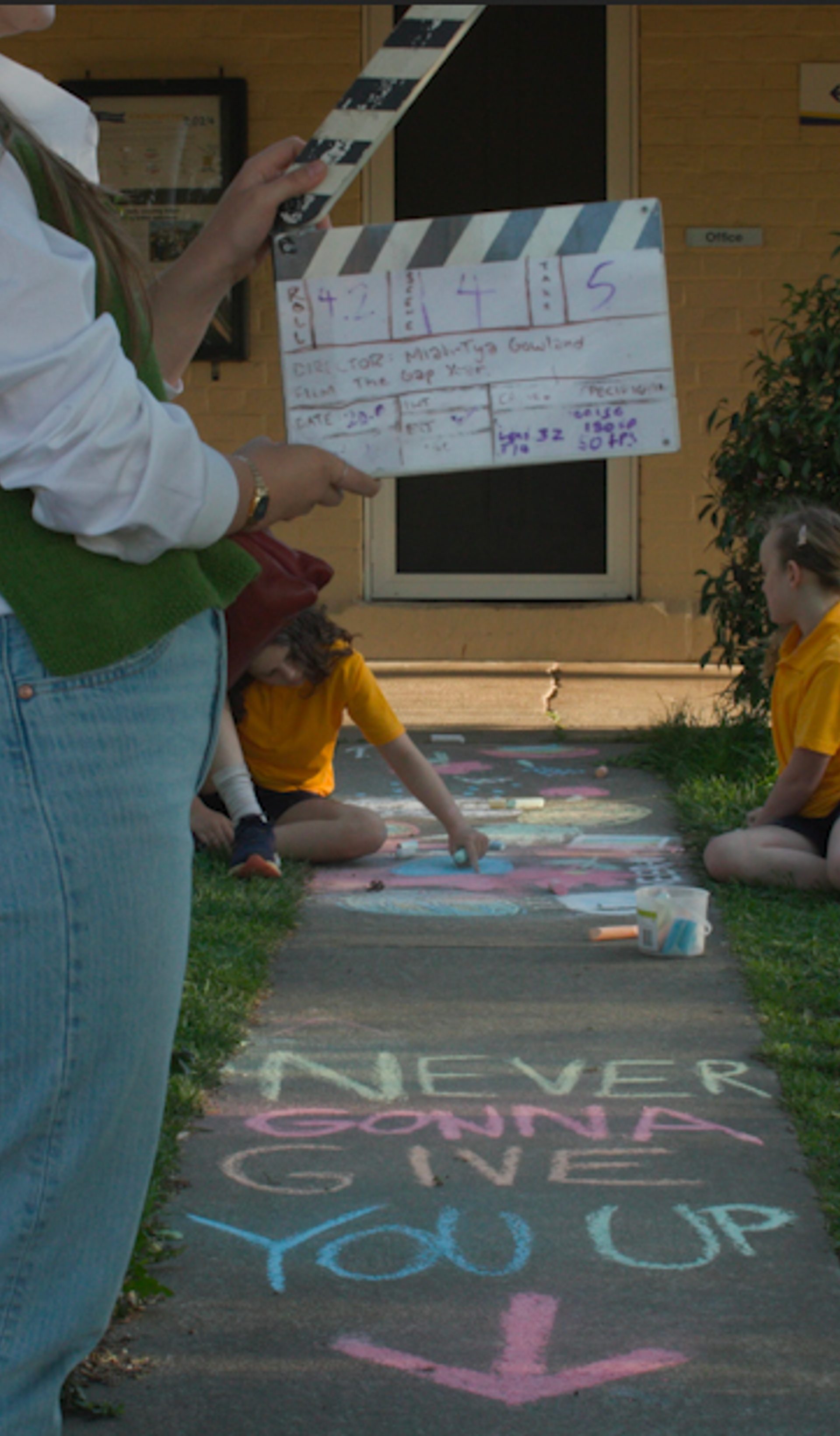 Crew member holding a clapper board with two young children in the background. Text written in chalk on the ground says "NEVER GONNA GIVE YOU UP"