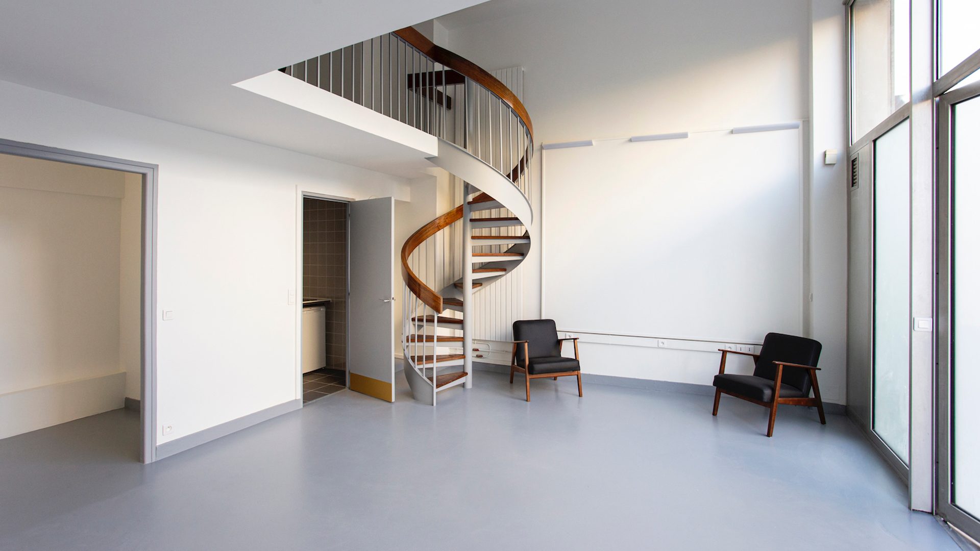 An open naturally lit room with a grey floor and white walls. There is a spiral staircase in the centre of the room and two grey arm chairs.