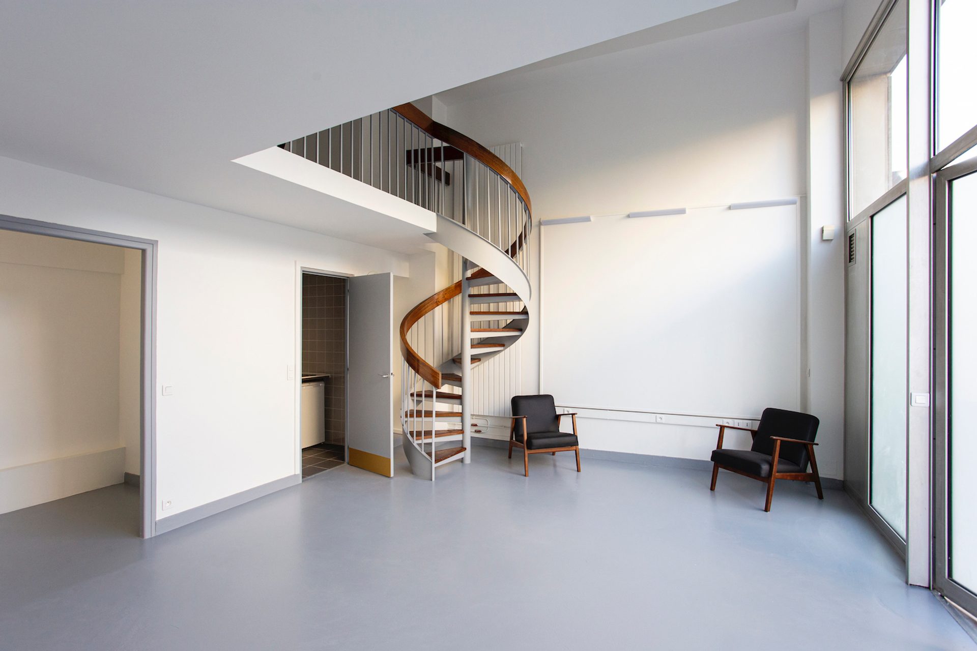 An open naturally lit room with a grey floor and white walls. There is a spiral staircase in the centre of the room and two grey arm chairs.