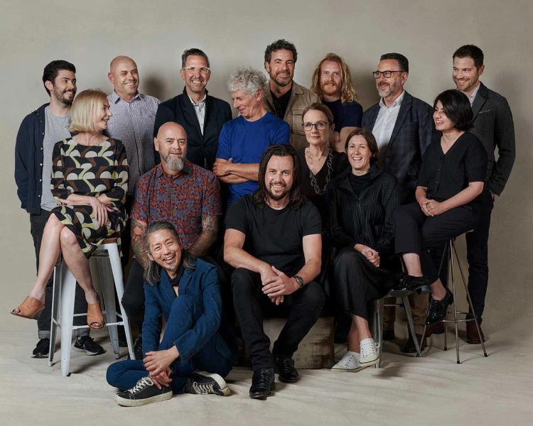 A group of people stand and sit on a beige background. Some look at the camera while others look candidly at each other.