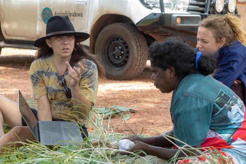Three women sitting outside on a woven green and yellow mat, which lies on red dirt.