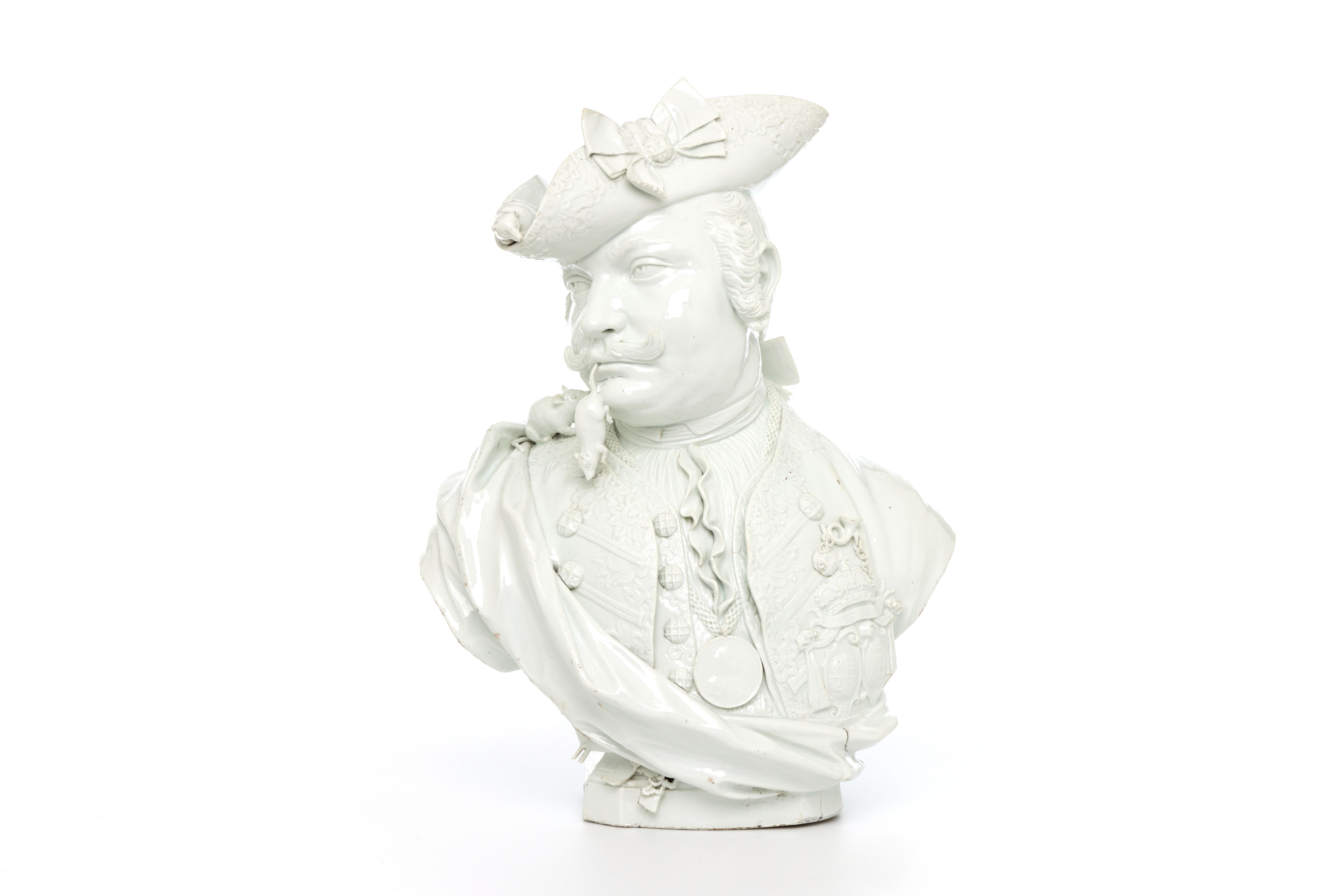 A head and shoulders satirical portrait bust in glazed white hard-paste porcelain. The piece is naturalistically modelled to portray a solidly built man with the head turned to his right, with an up curled moustache and wearing a wig with queue (pigtail) wound around with a ribbon.