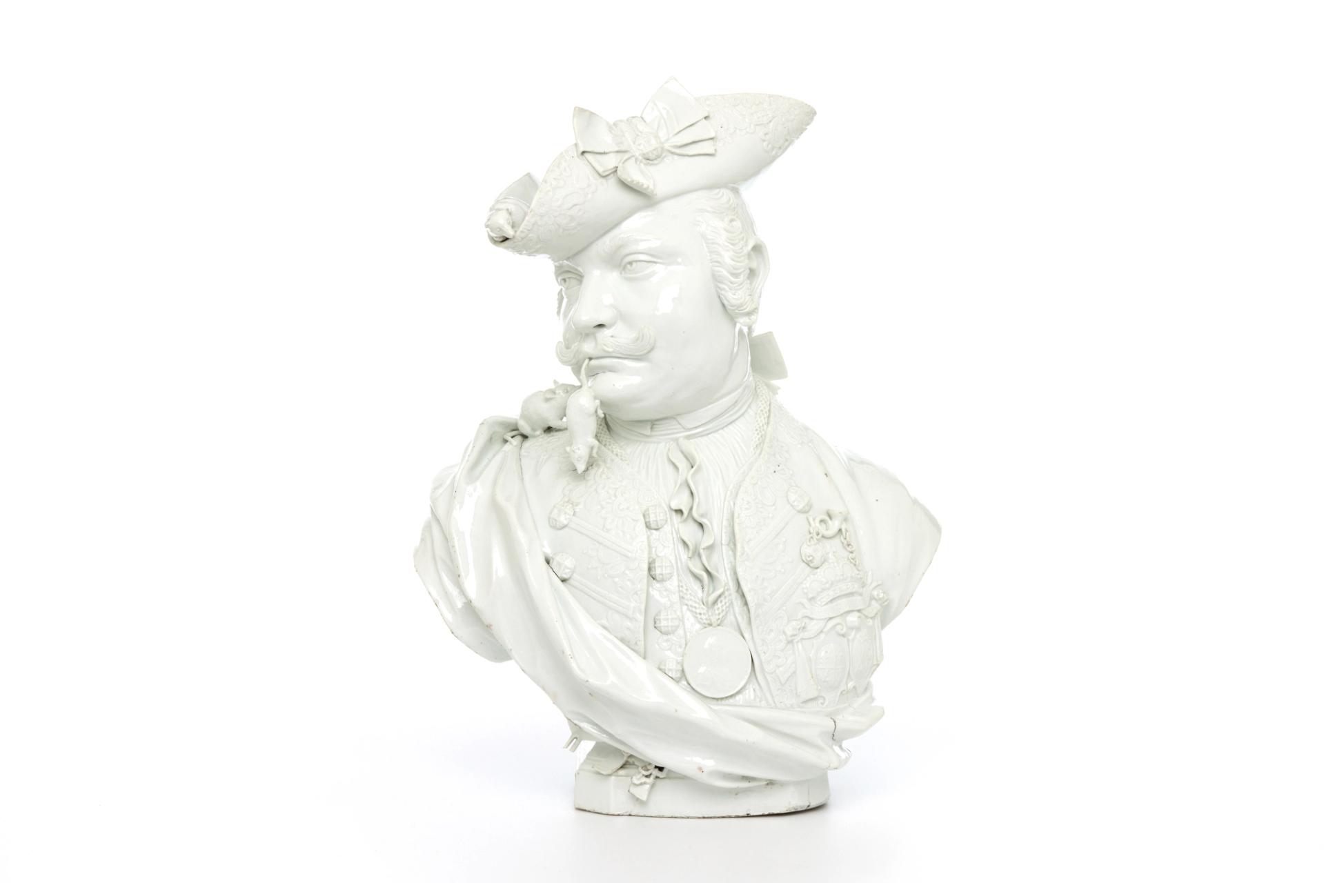A head and shoulders satirical portrait bust in glazed white hard-paste porcelain. The piece is naturalistically modelled to portray a solidly built man with the head turned to his right, with an up curled moustache and wearing a wig with queue (pigtail) wound around with a ribbon.