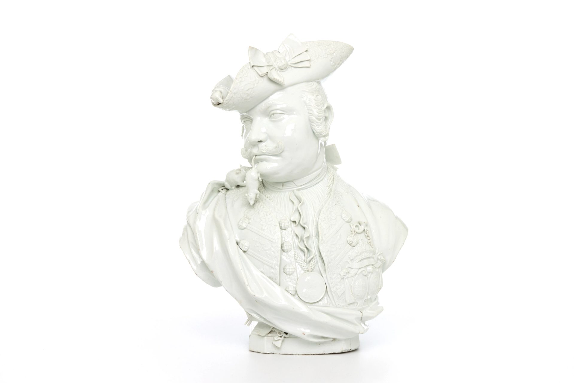 A head and shoulders satirical portrait bust in glazed white hard-paste porcelain. The piece is naturalistically modelled to portray a solidly built man with the head turned to his right, with an up curled moustache and wearing a wig with queue (pigtail) wound around with a ribbon.