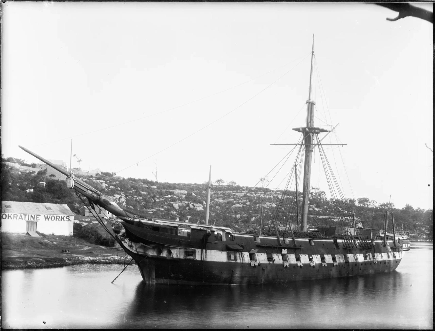 Glass plate negative depicting a coaling hulk, formerly the warship HMS 'Nelson' moored near Neokratine Explosive Co.'s works, Kerosene Bay, Sydney. Landscape-format, black and white glass plate negative depicting a coaling hulk, converted from an old warship with gun ports. The background features a small shed on the left hand side with the text 'Okratine Works' on the side.