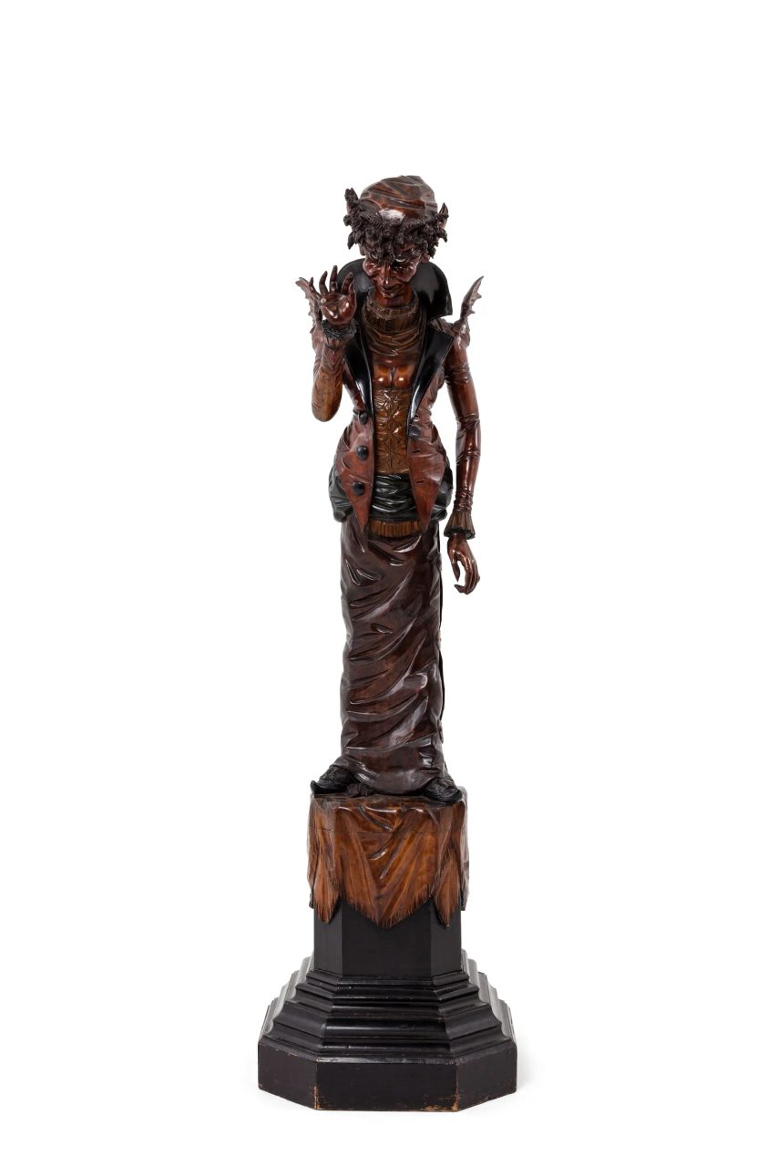 Carved pine wood figure of a standing female, faun-like being dressed in an interpretation of late 1800s women's fashion standing on a 600mm high stepped octagonal pedestal, the upper section with carved drapery. On its pedestal, the figure stands almost two metres tall and has an exaggerated S-bend posture which pushes the head forward and buttocks out.