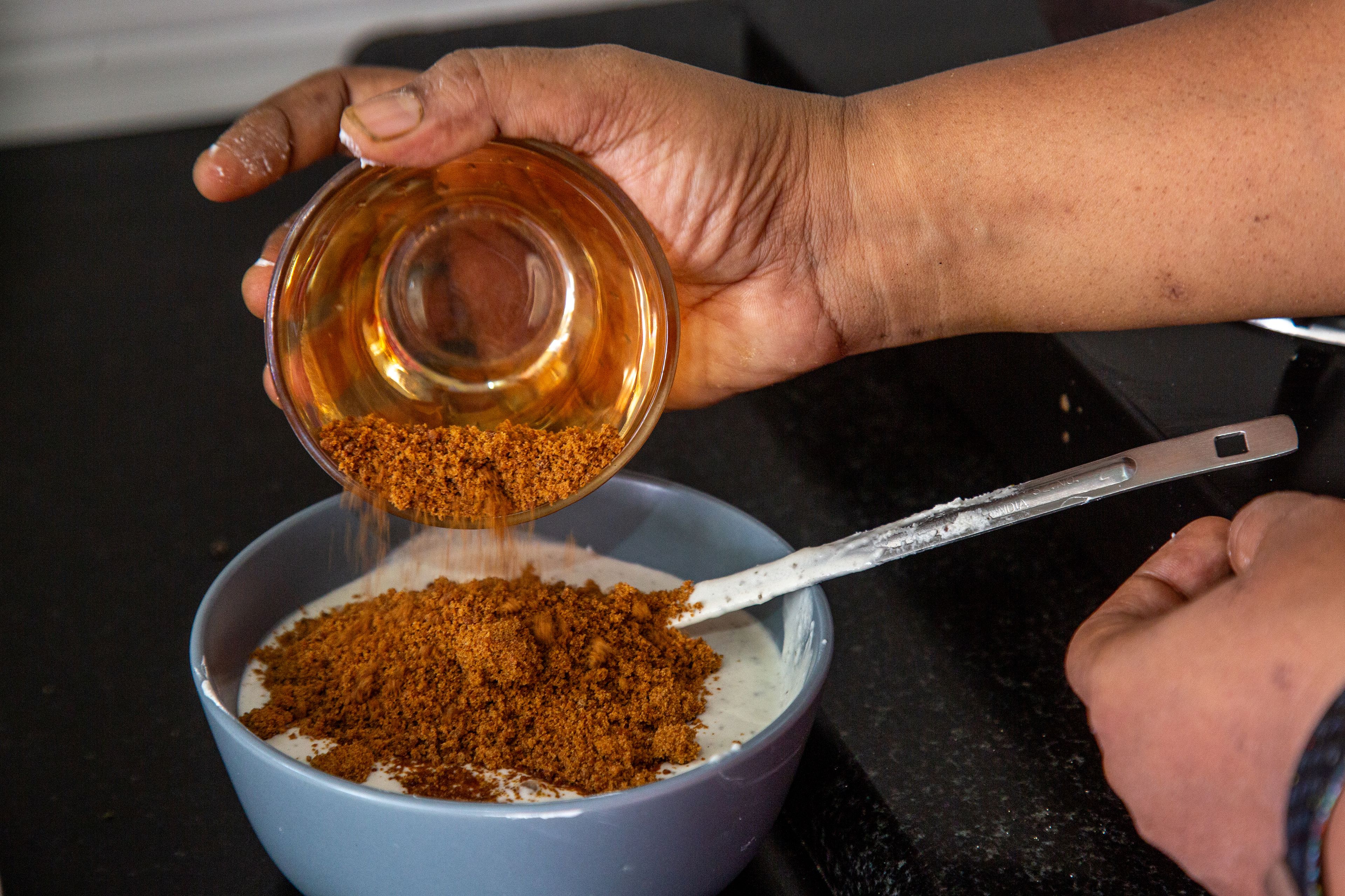 A brown hand tips a bowl of sugar made from the sap of palms into a bowl of condensed milk.