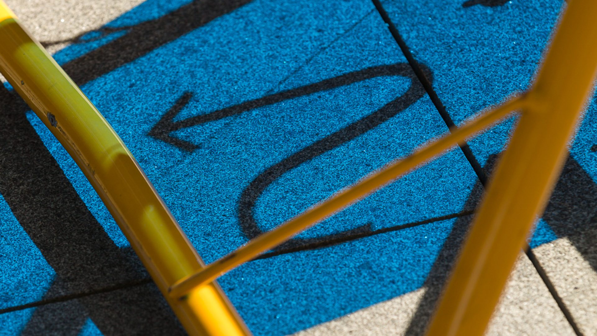 A blue coloured shadow depicting a curvy arrow cast on to concrete, with a yellow tubular steel frame in the foreground
