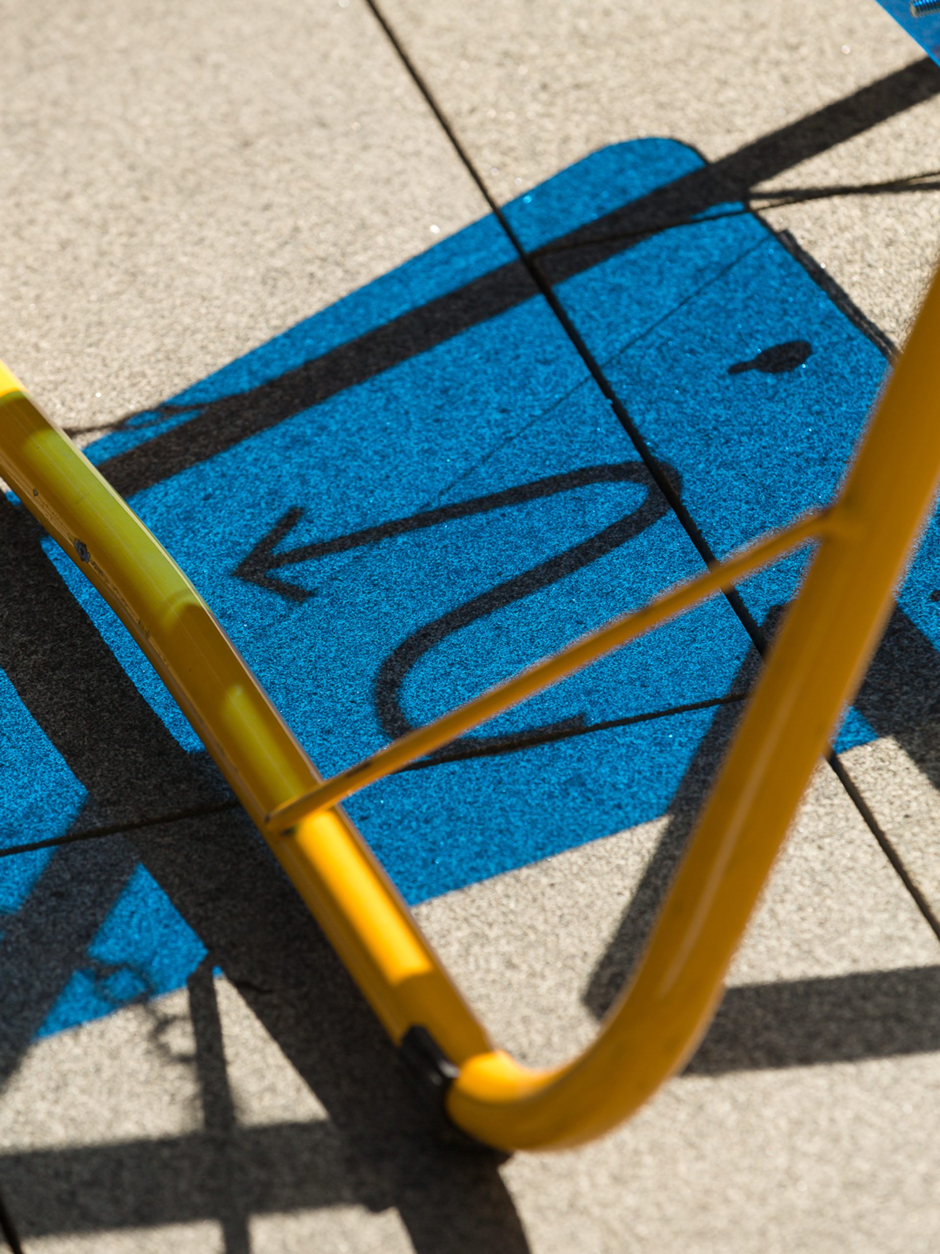 A blue coloured shadow depicting a curvy arrow cast on to concrete, with a yellow tubular steel frame in the foreground