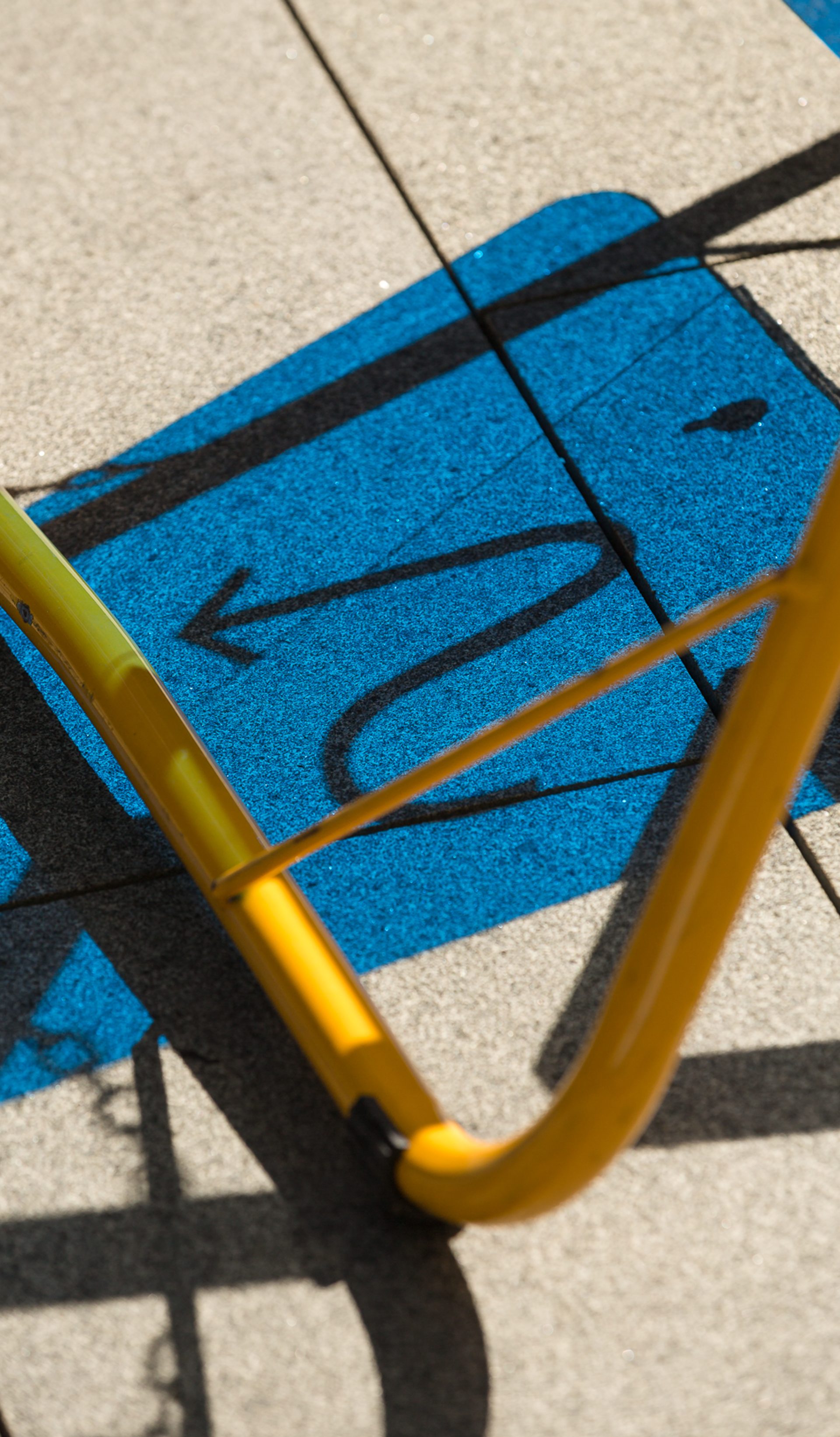 A blue coloured shadow depicting a curvy arrow cast on to concrete, with a yellow tubular steel frame in the foreground