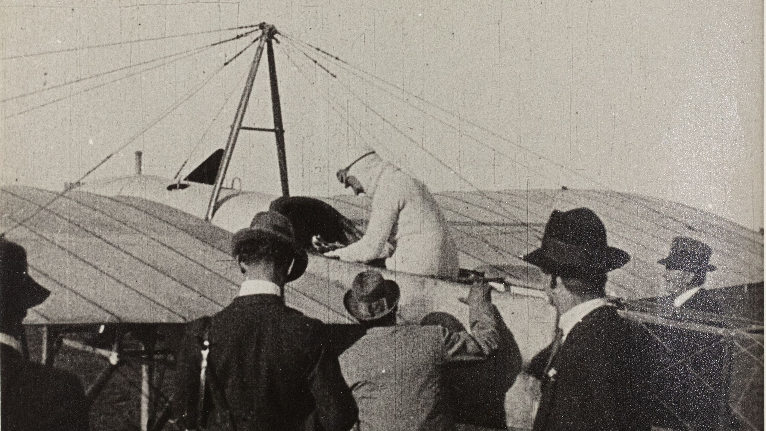 Black and white photograph of the first Australian airmail flight at Government House Melbourne. Multiple men in suits and hats looking to a pilot sitting on a plane, preparing to fly.