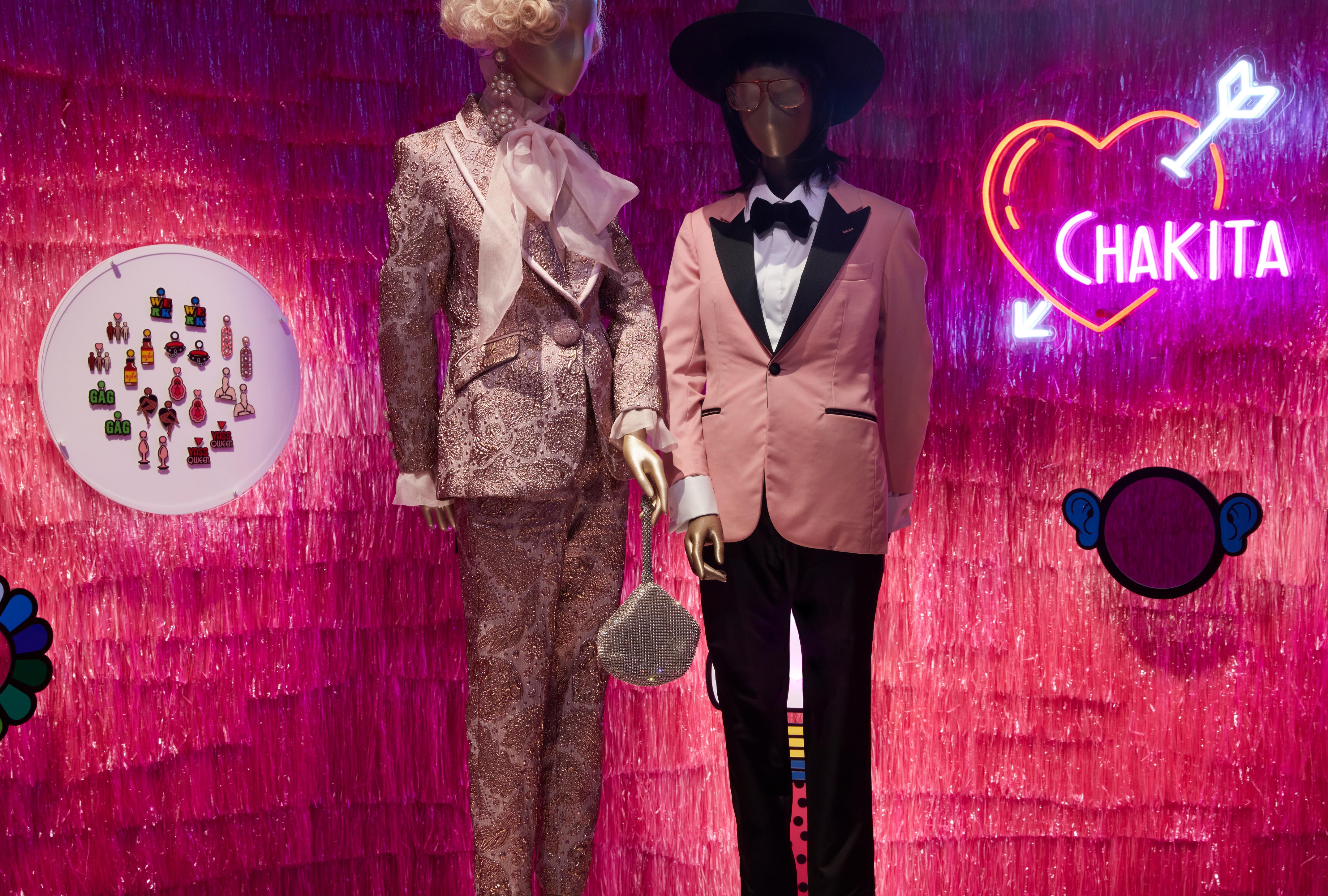 Two mannequins wearing pink suits and accessories standing in front of a wall lined with pink glitter streamers. A neon light love heart with an arrow through it and ‘CHAKITA’ hangs on the wall.