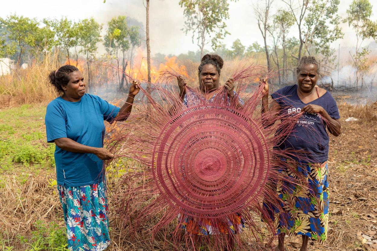 Three women holding a large pink traditional weaving. They are outside and a fire is visible in the background.