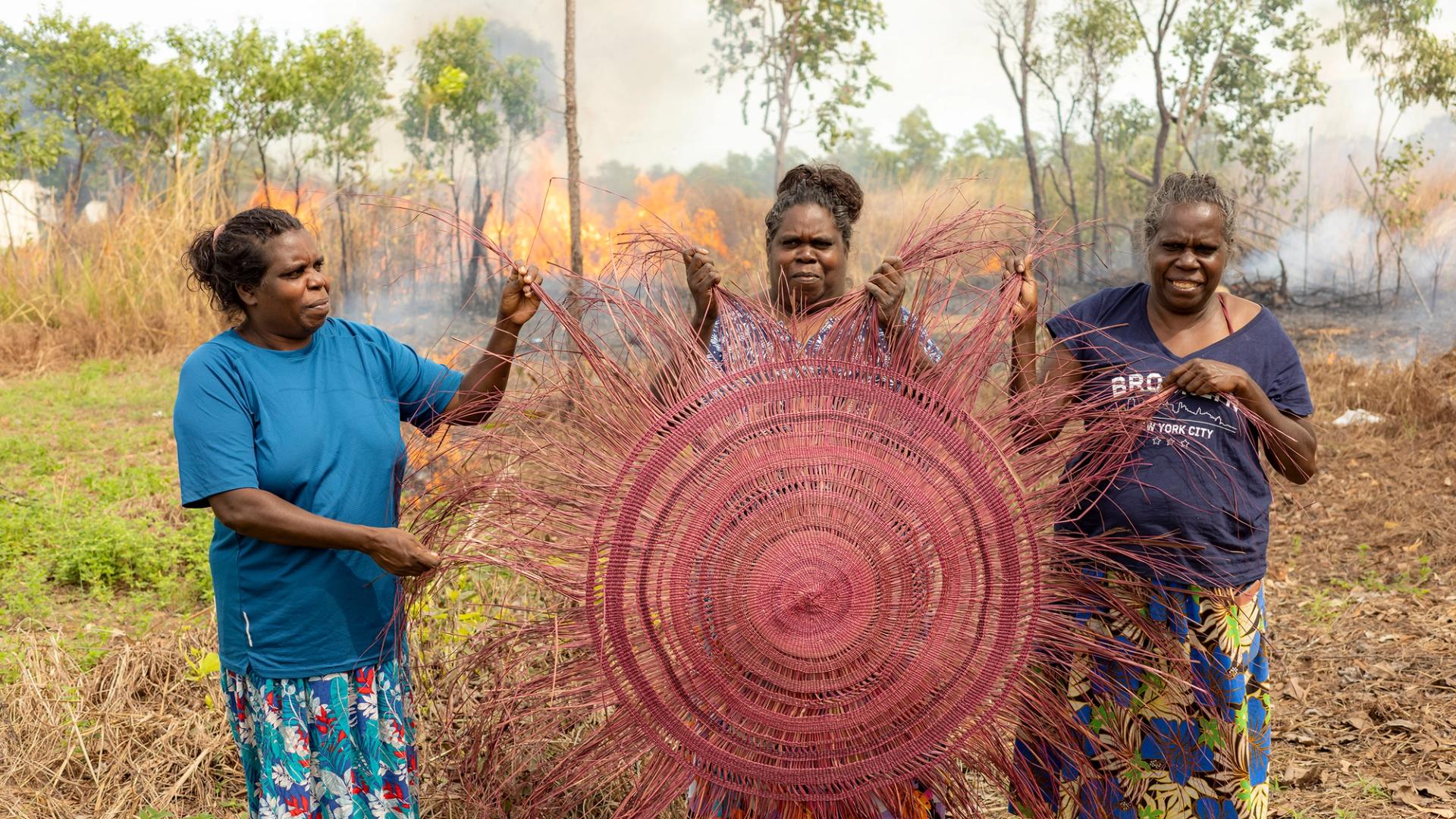 Three women holding a large pink traditional weaving. They are outside and a fire is visible in the background.