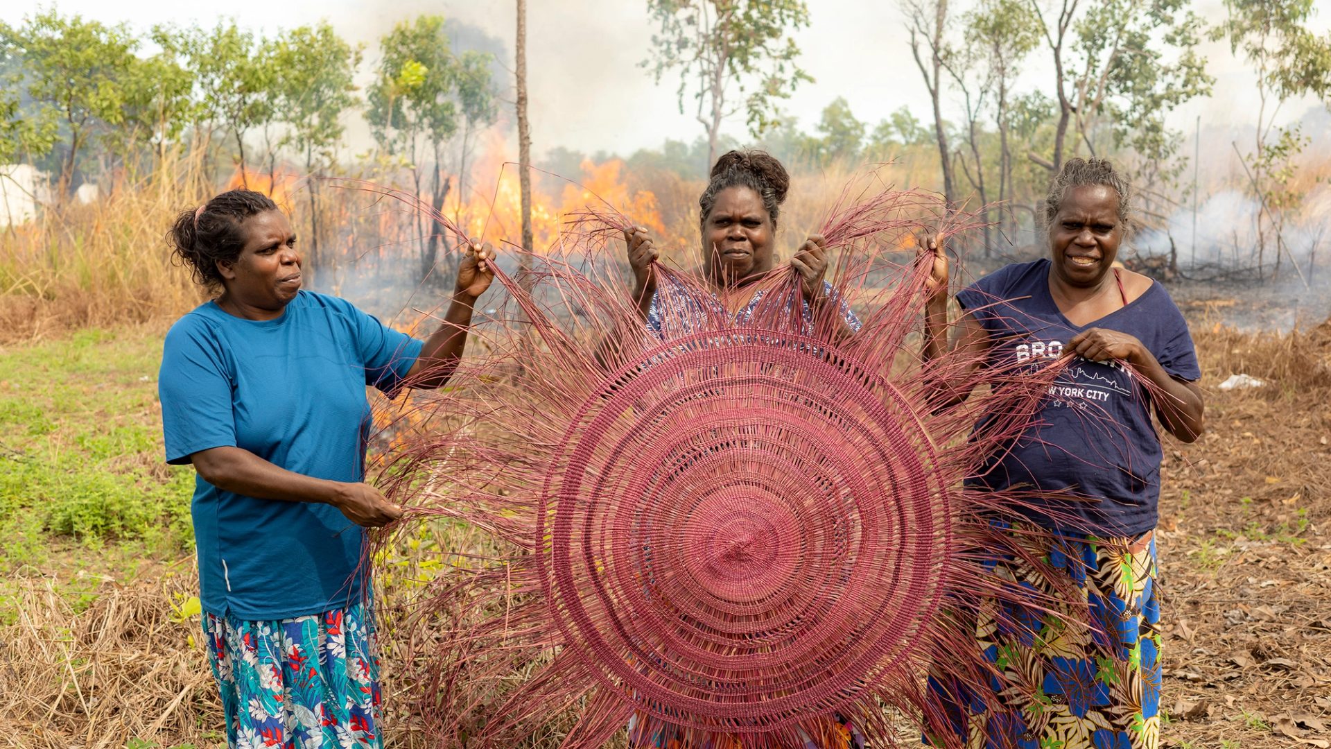Three women holding a large pink traditional weaving. They are outside and a fire is visible in the background.
