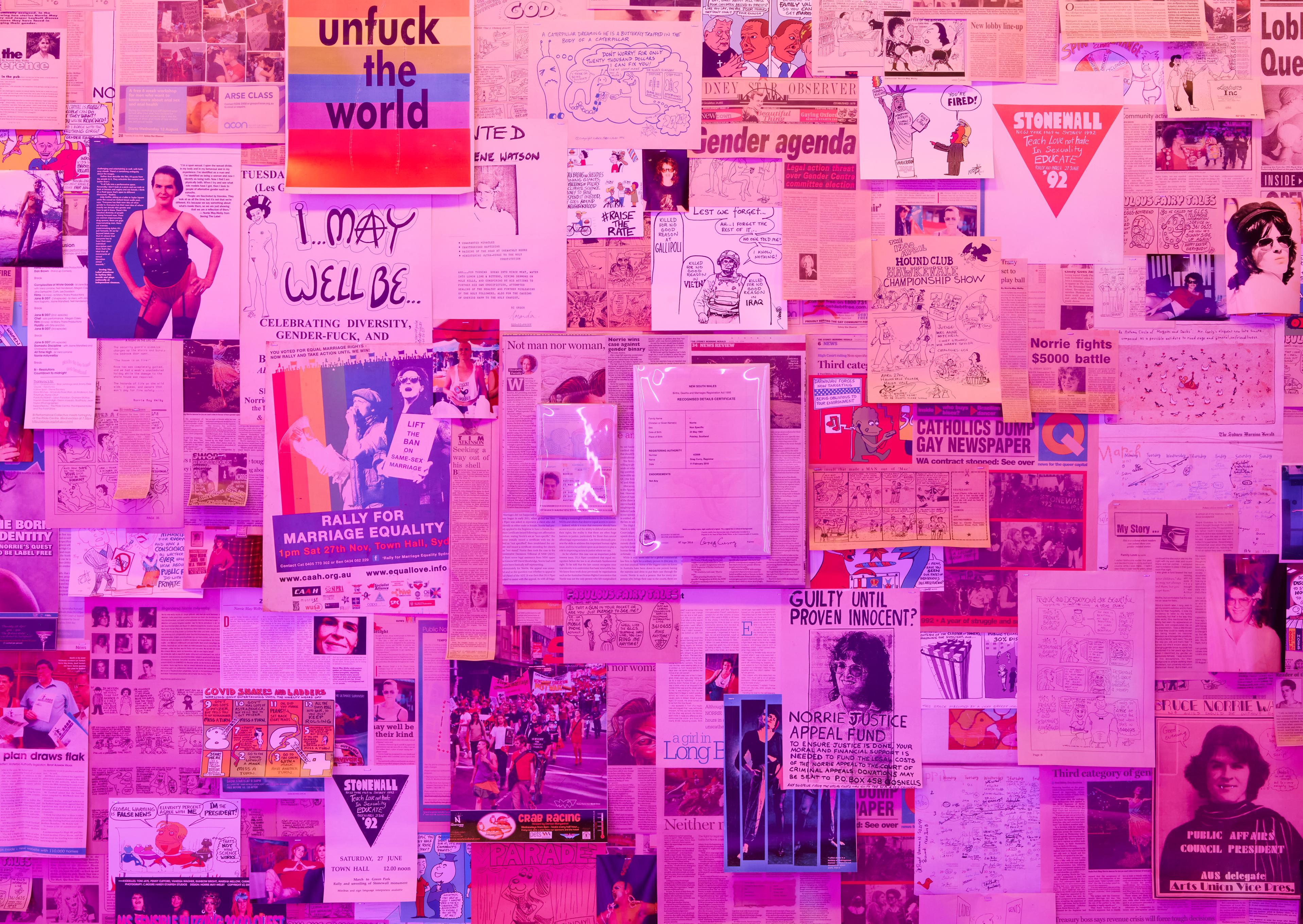 A wall with images, newspaper clippings, and posters collaged together cast with pink light. Mounted on the wall are two documents, a passport and birth certificate, under plexiglas.