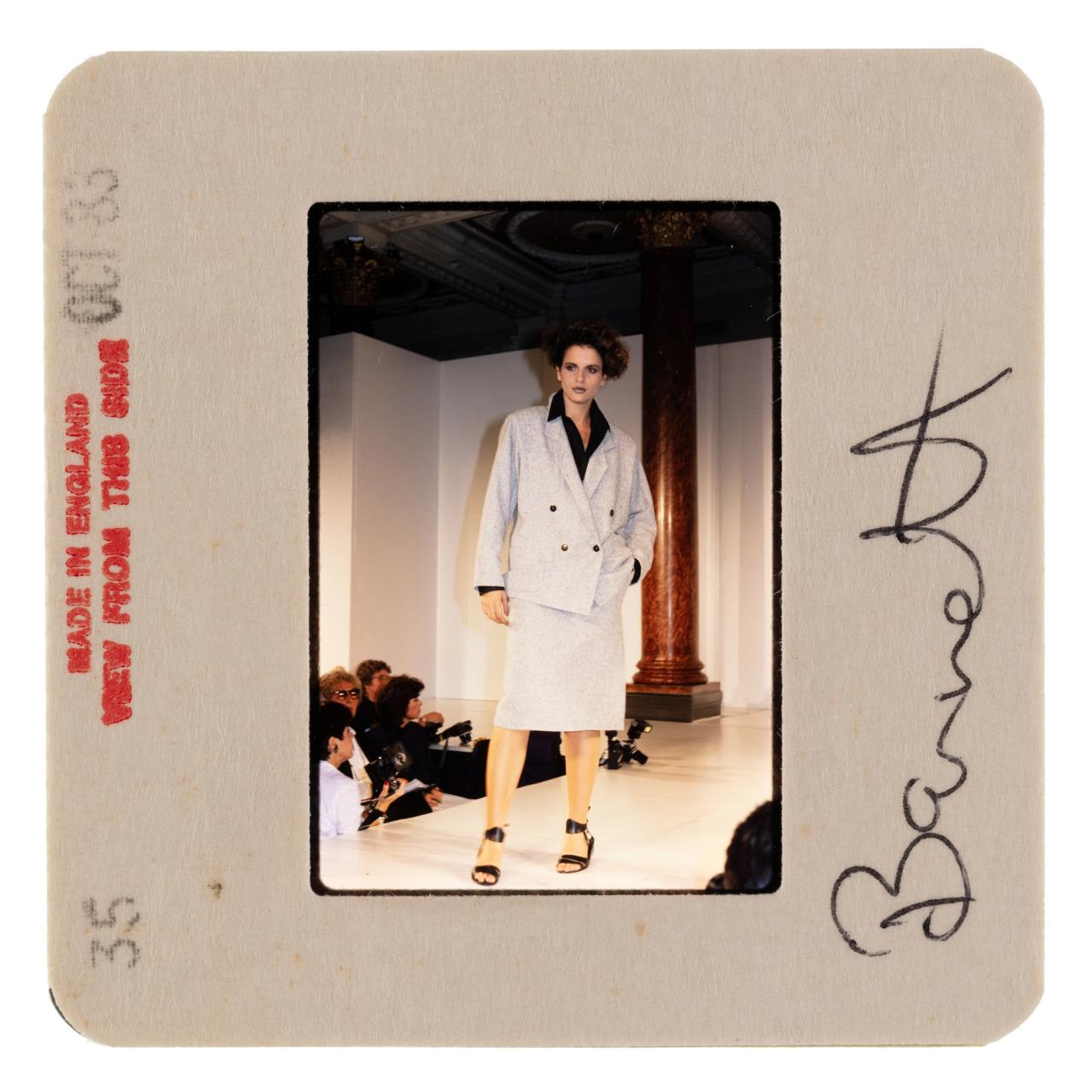 A photo positive 35mm slide, depicting a fashion show and runway model wearing a light grey blazer suit jacket and midi length pencil skirt with black buttons, undercollar shirt and heels.