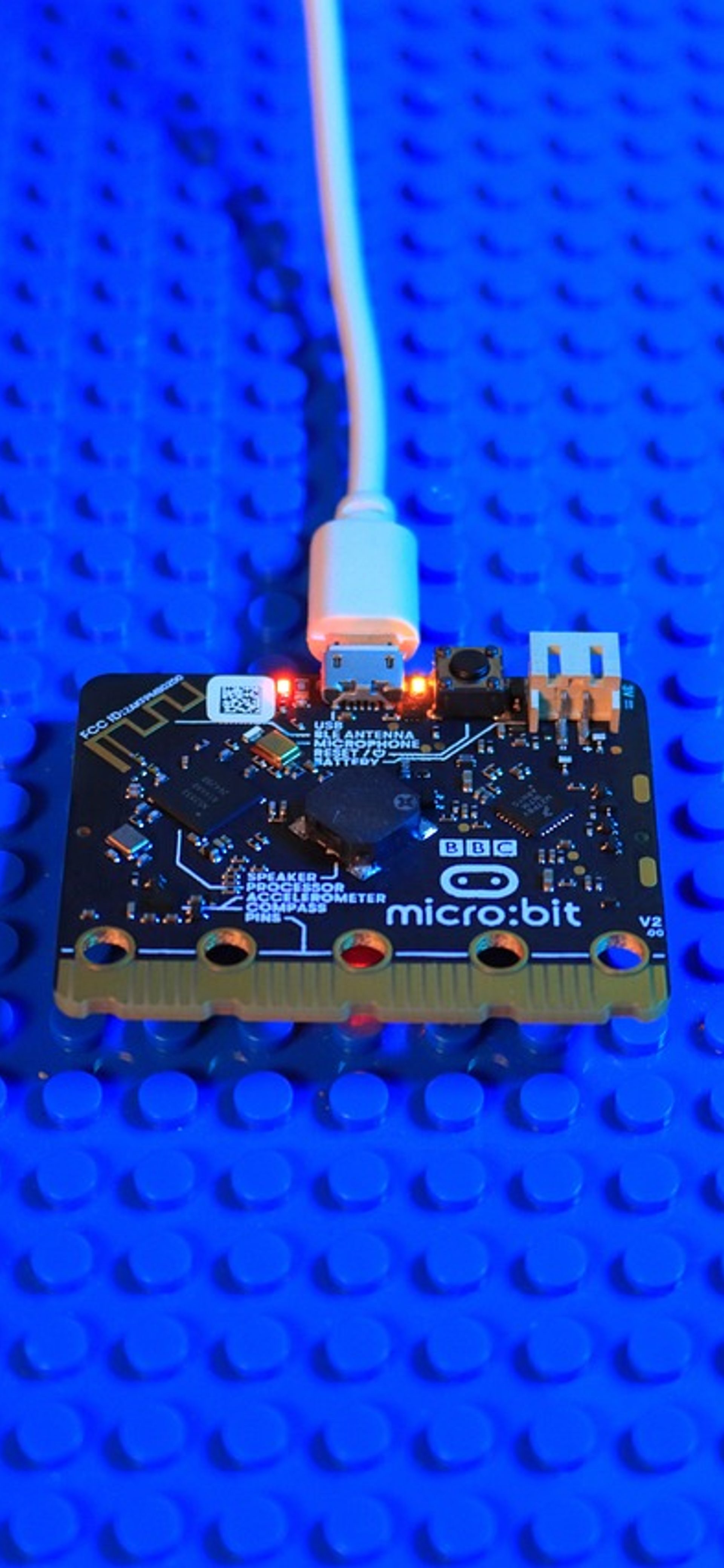 A black and copper BBC micro:bit microprocessor with a white connecting cable sits on a blue Lego base plate.