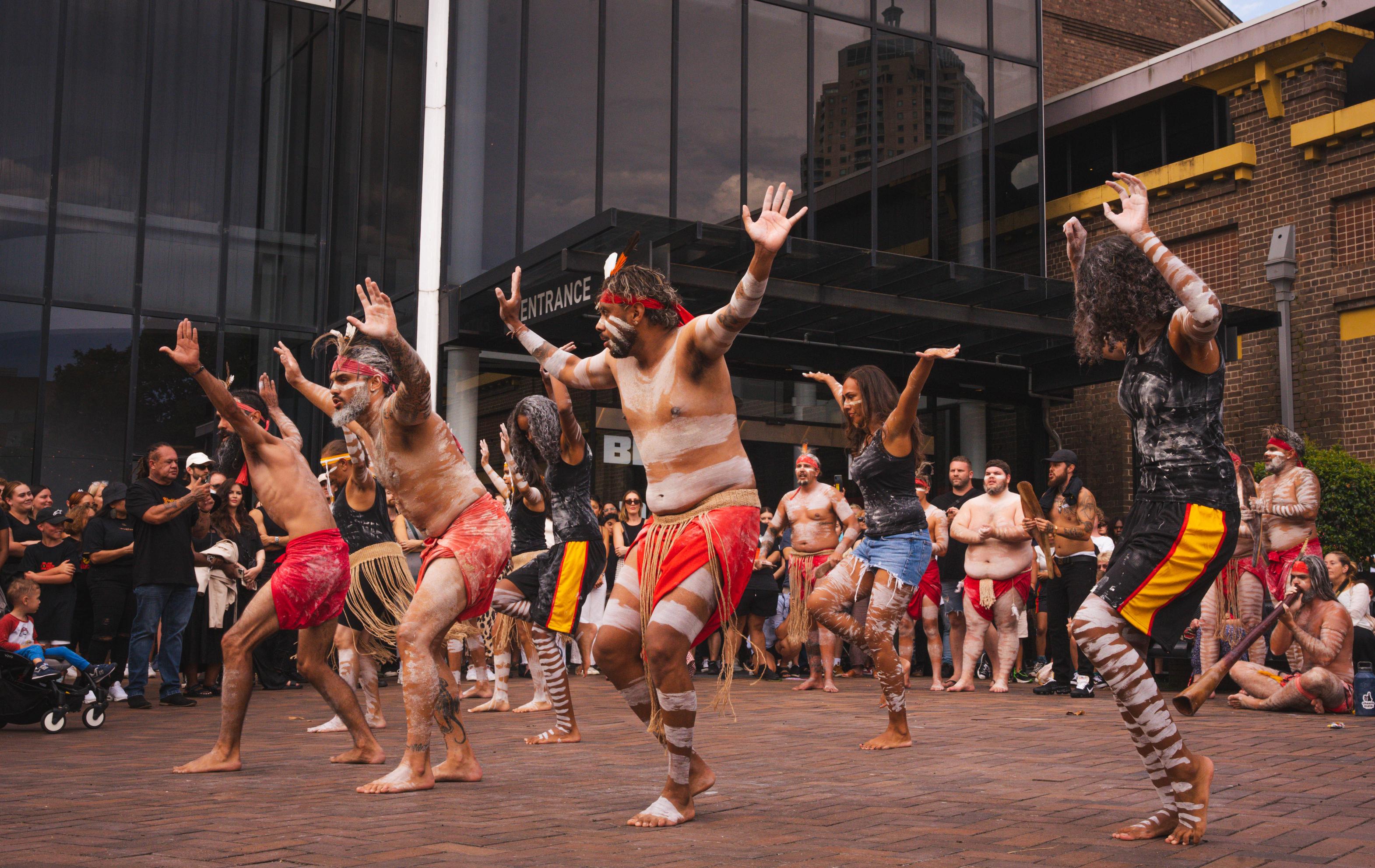 A group of First Nations performers in traditional dress, mid dance. They are outside with an audience watching them.