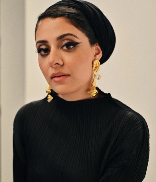 Female figure looking at the camera wearing artistic gold faces as earings, eyeliner and all black.