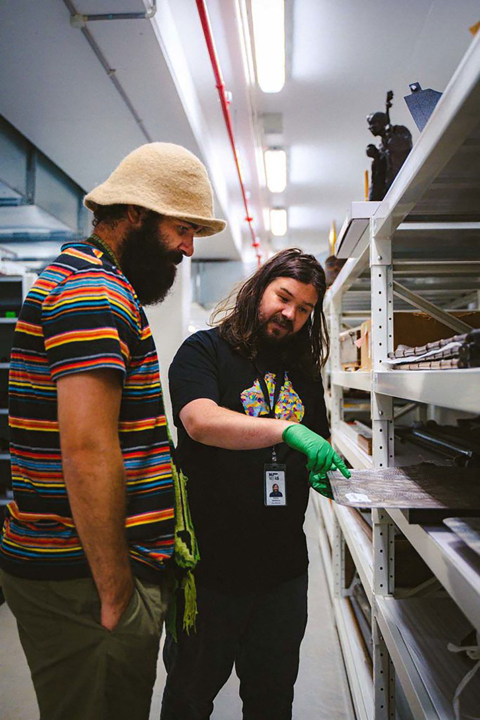 Joseph Douglas with a Powerhouse staff member observing a painting from the collection. The staffer is wearing green rubber gloves while pulling the painting off a metal shelf and pointing to a specific part of the image.