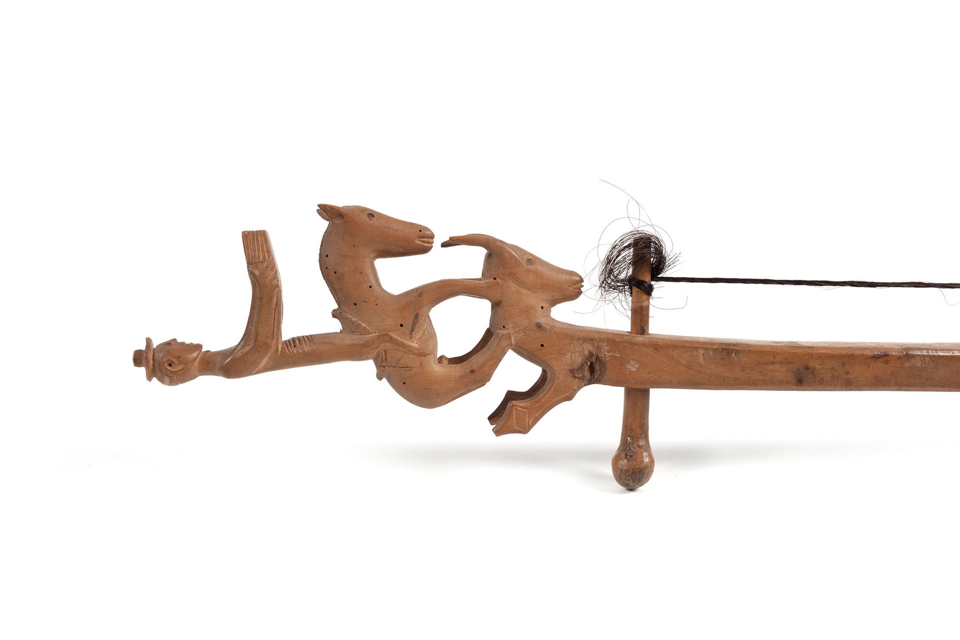 A carved wooden ornament of a man standing on a donkey which is standing on the head of a horned goat head at the end of a wooden stringed instrument.