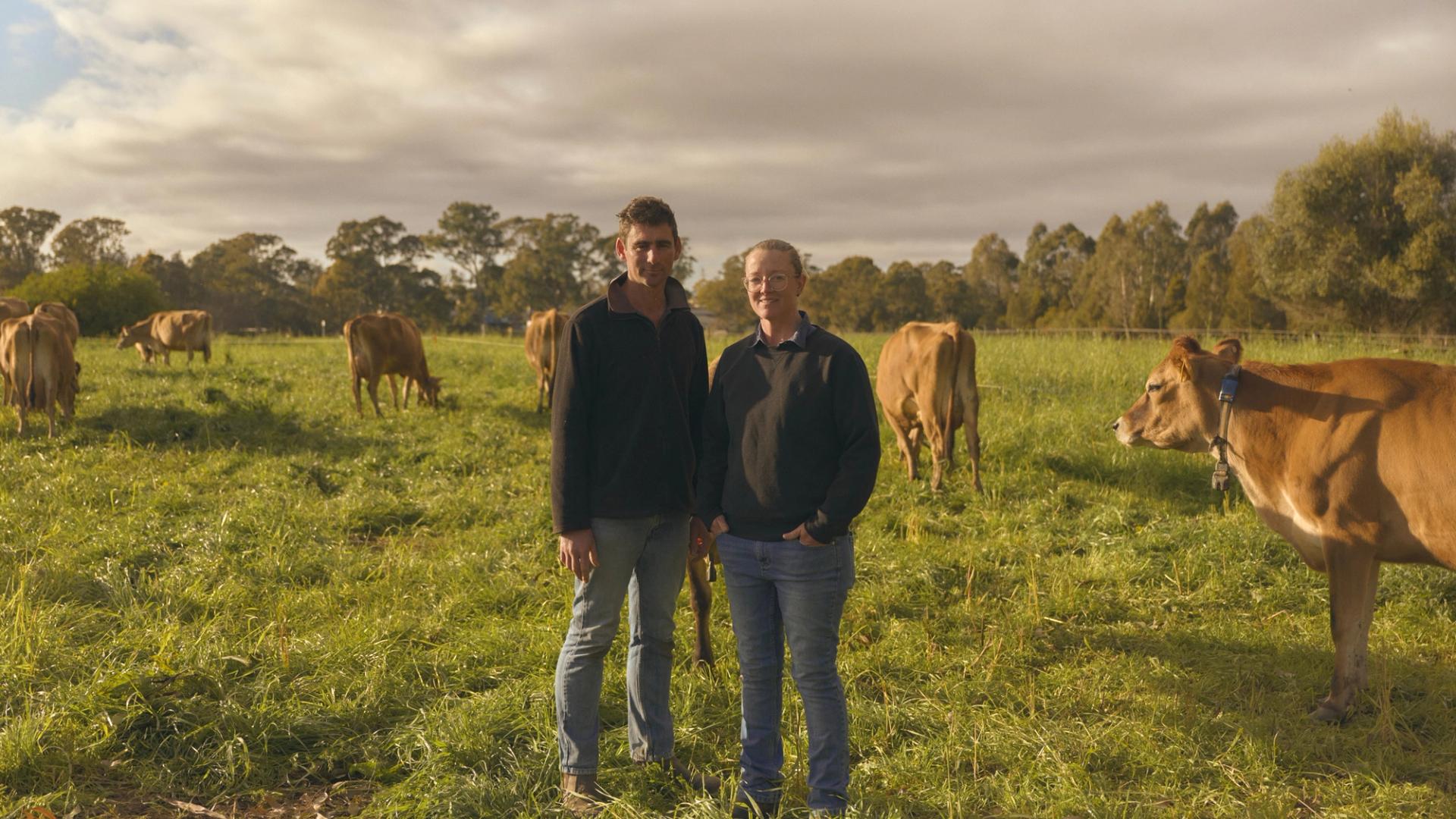 Two farmers stand side by side in a lush green pasture, surrounded by grazing cows. Both are dressed in casual outdoor clothing, with the man on the left wearing a dark jacket and jeans, and the woman on the right in a similar outfit. The cows, with their light brown coats, are calmly eating grass, and the scene is set under a partly cloudy sky, with trees visible in the background.