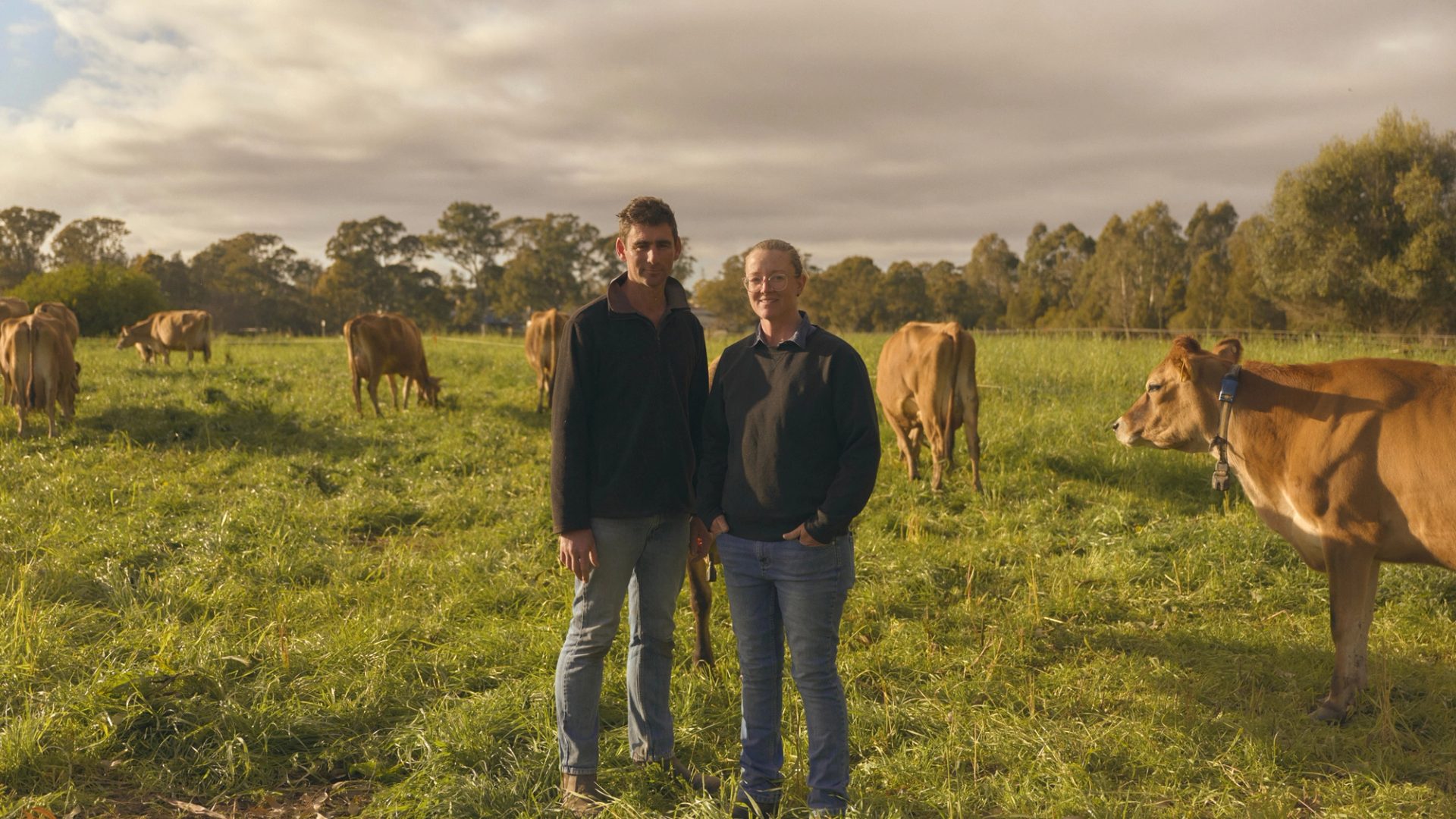 Two farmers stand side by side in a lush green pasture, surrounded by grazing cows. Both are dressed in casual outdoor clothing, with the man on the left wearing a dark jacket and jeans, and the woman on the right in a similar outfit. The cows, with their light brown coats, are calmly eating grass, and the scene is set under a partly cloudy sky, with trees visible in the background.