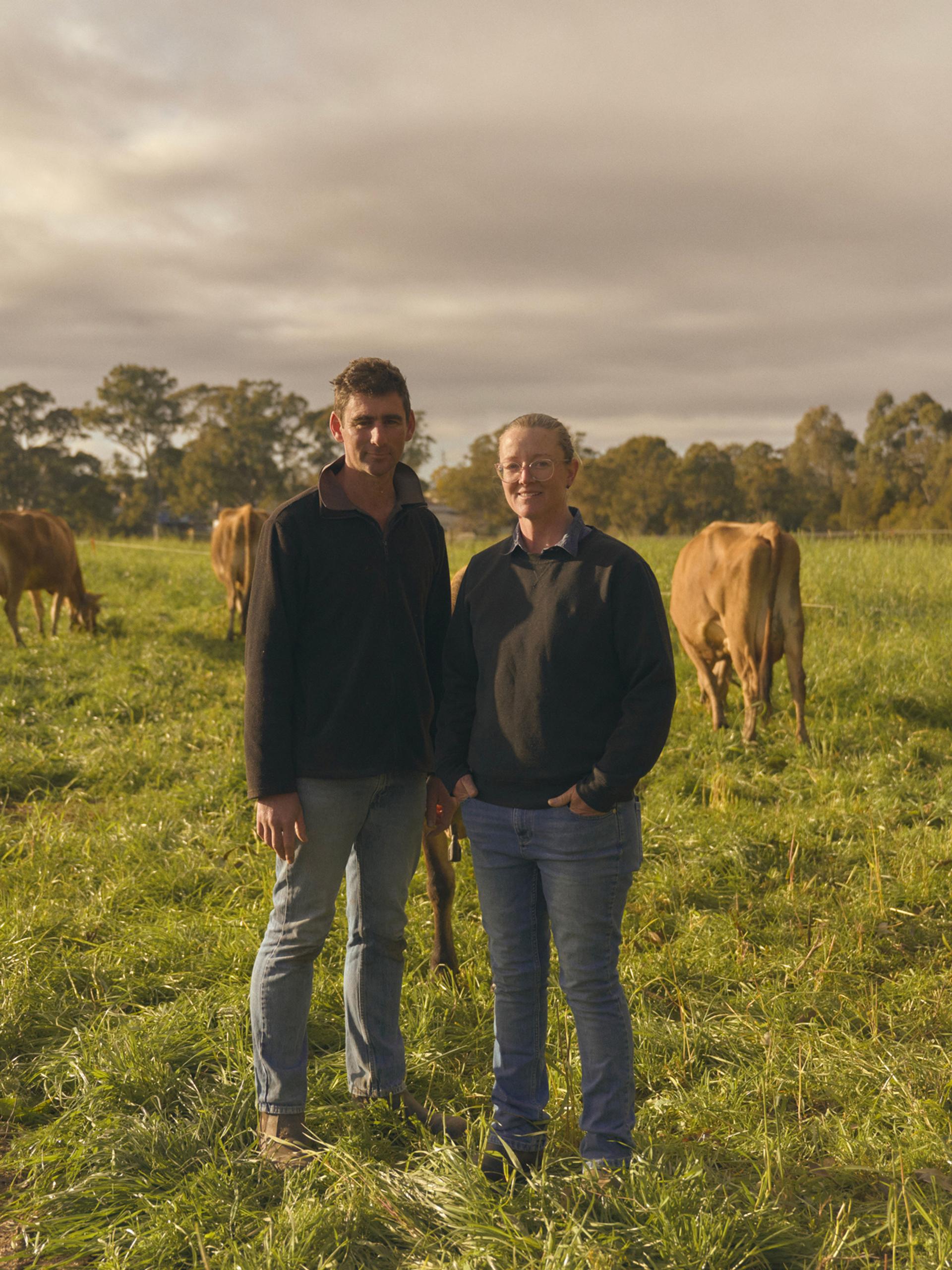 Two farmers stand side by side in a lush green pasture, surrounded by grazing cows. Both are dressed in casual outdoor clothing, with the man on the left wearing a dark jacket and jeans, and the woman on the right in a similar outfit. The cows, with their light brown coats, are calmly eating grass, and the scene is set under a partly cloudy sky, with trees visible in the background.